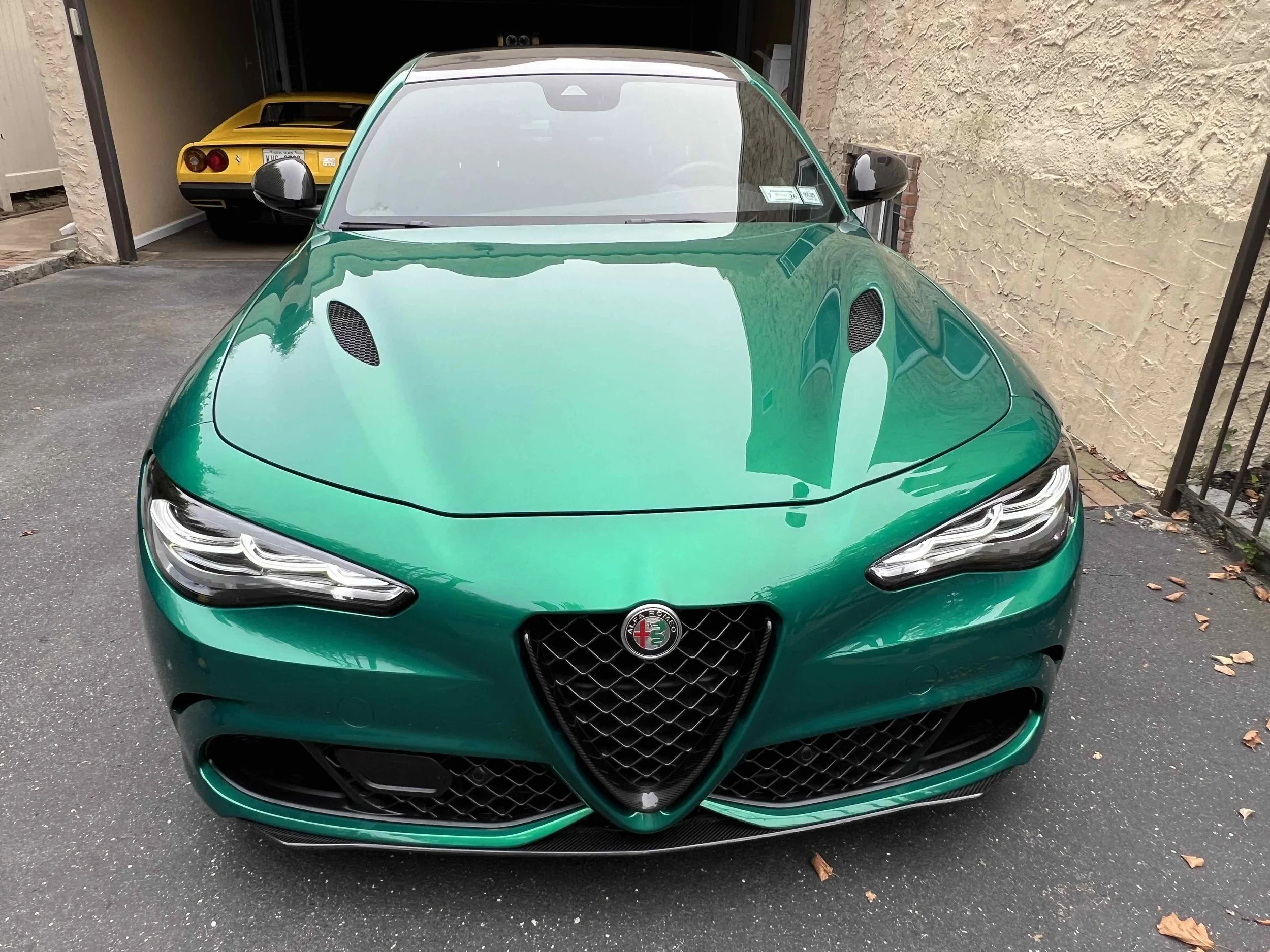 Front view of a green Alfa Romeo sports car parked in a driveway with a yellow sports car visible in the garage behind it.
