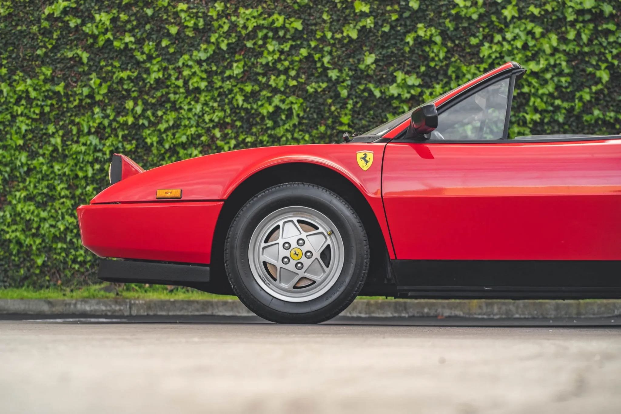 A red vintage Ferrari sports car parked on a street with a green hedge in the background.