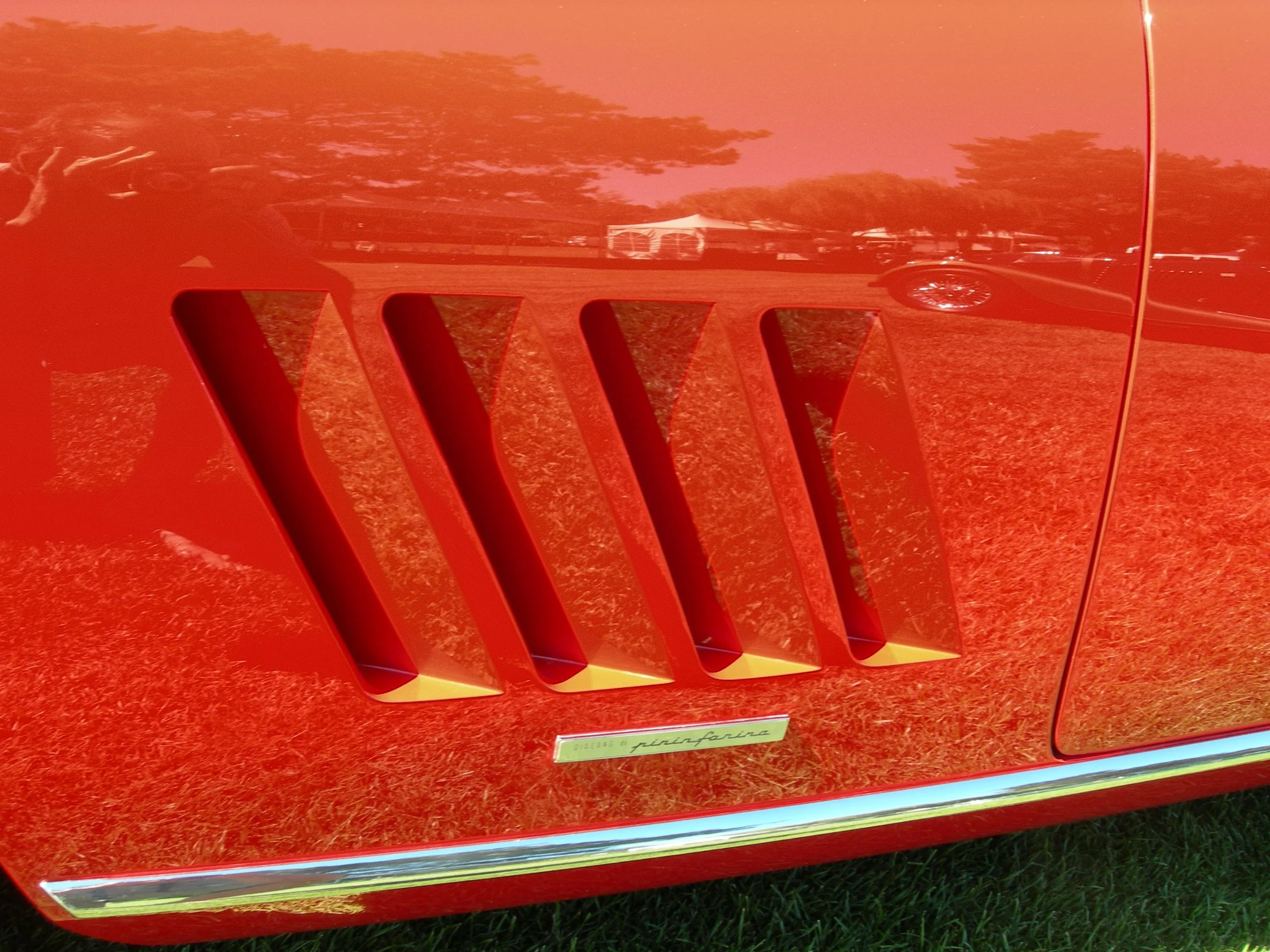 Close-up of a red Ferrari with four side vents and a small Ferrari emblem below them. Reflects trees and sky on its glossy surface.