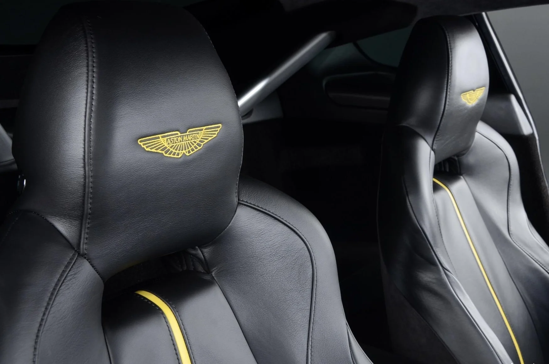 Close-up of two black leather car seats with yellow embroidered Aston Martin logos on the headrests and yellow piping, inside a luxury vehicle.