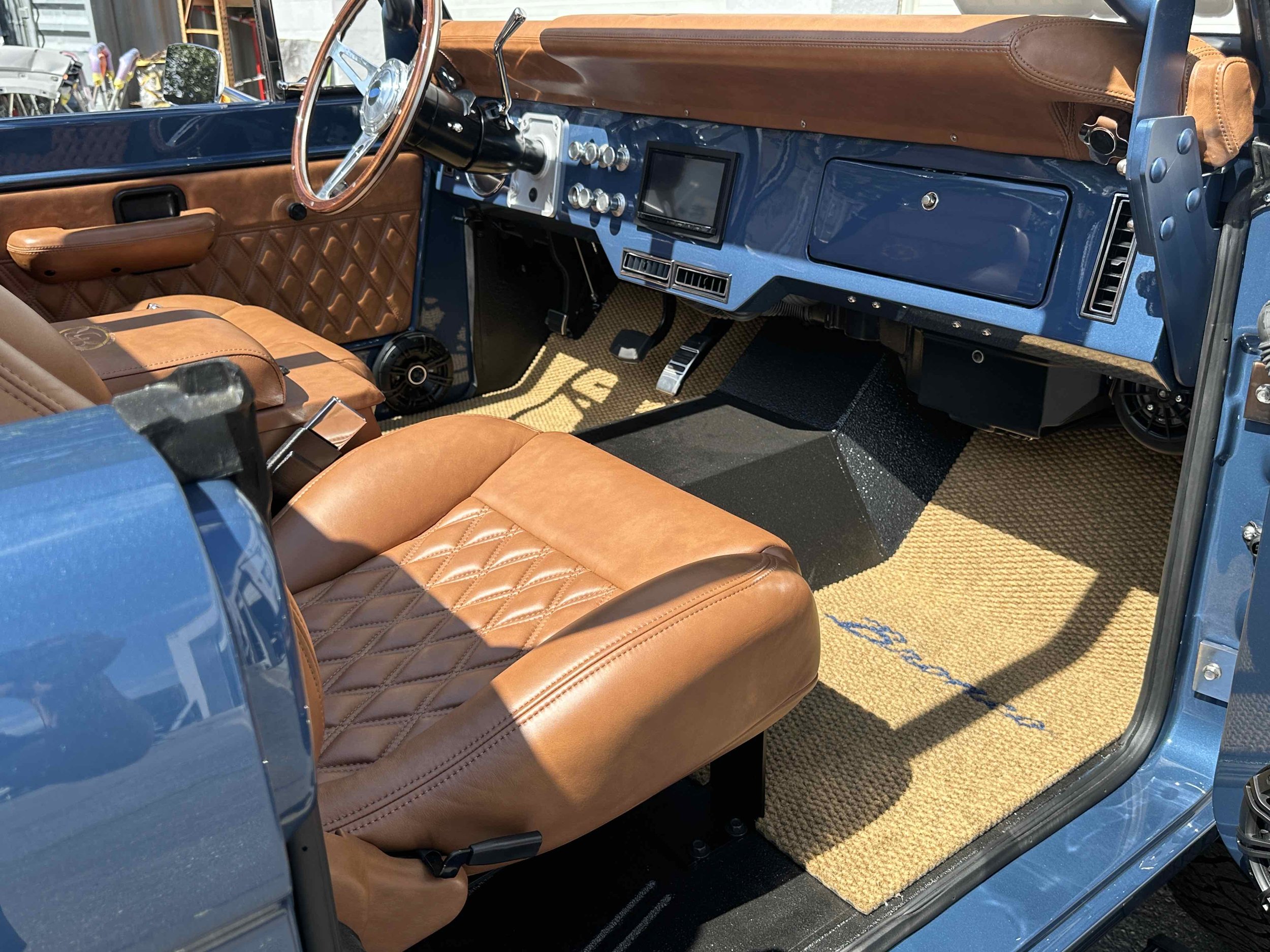 Inside of a vintage car with tan leather seats, a blue dashboard, and tan floor mats. The steering wheel and dashboard controls are visible, with the driver’s seat and interior panels also in tan leather. There are black and blue accents throughout t
