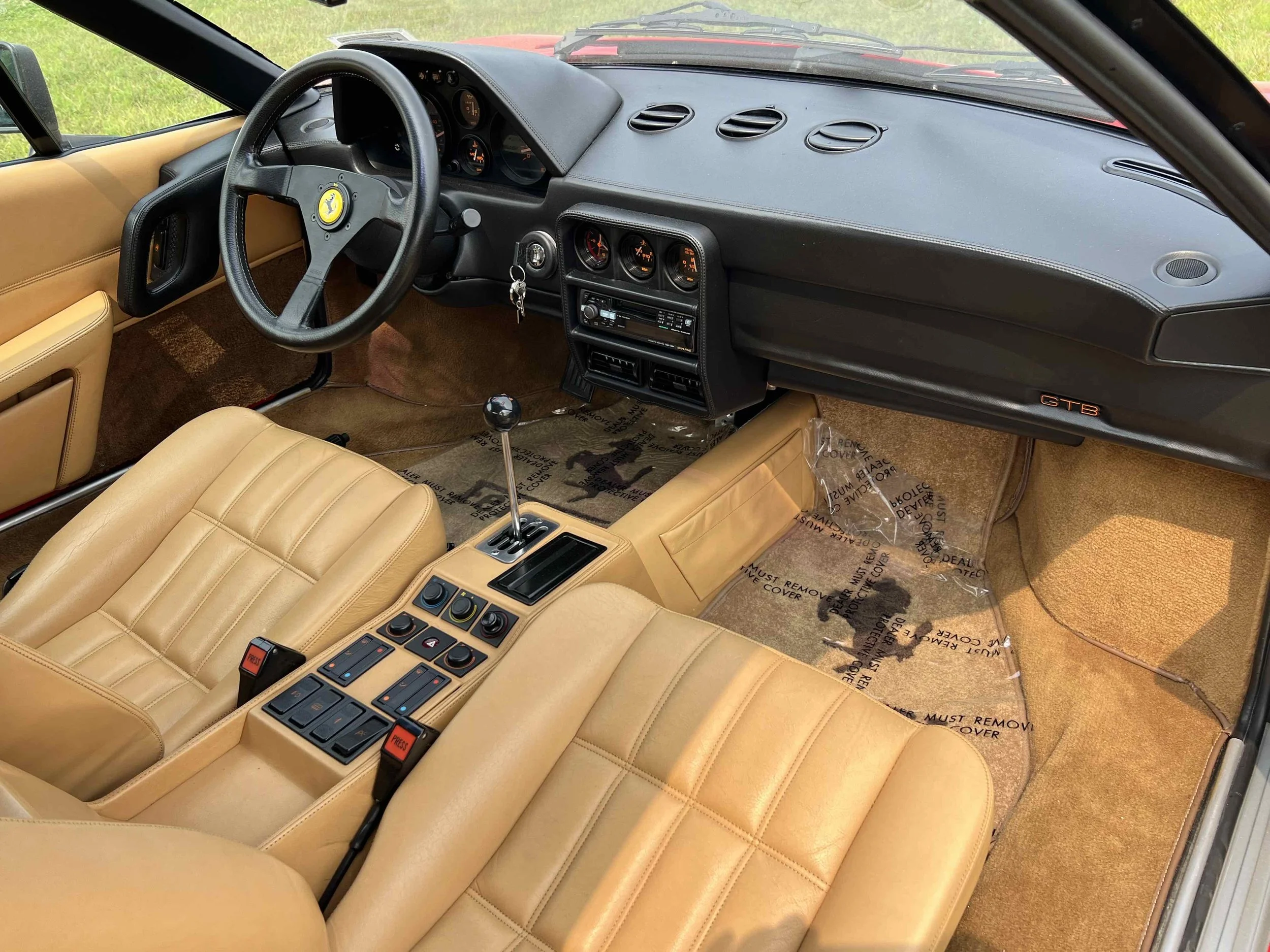 Interior of a vintage Ferrari GTB with tan leather seats, black steering wheel with Ferrari emblem, classic dashboard gauges, center console with gear shift and multimedia controls.