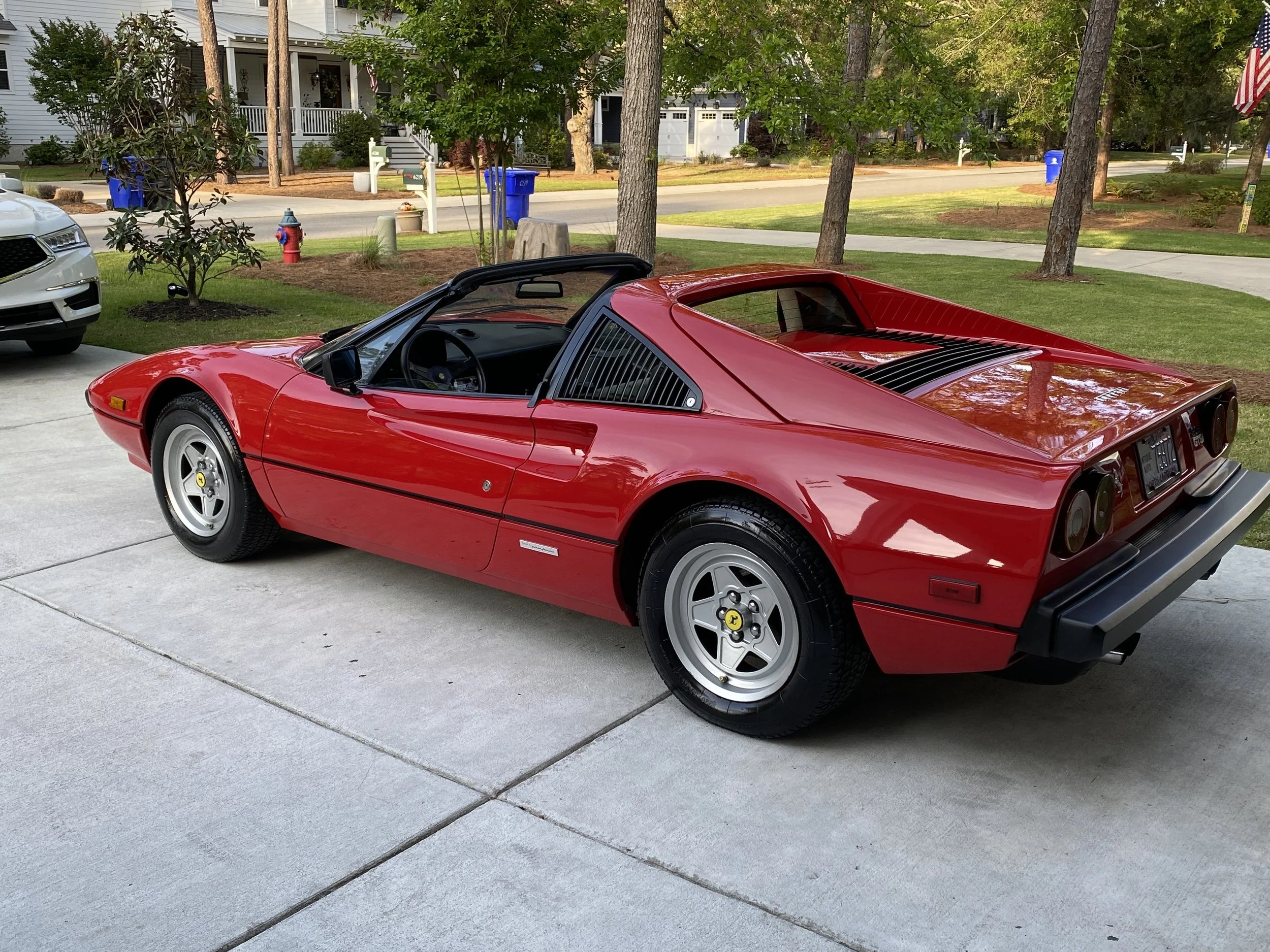 Red vintage Ferrari sports car parked on a concrete driveway in a residential neighborhood, with a white car and houses visible in the background.