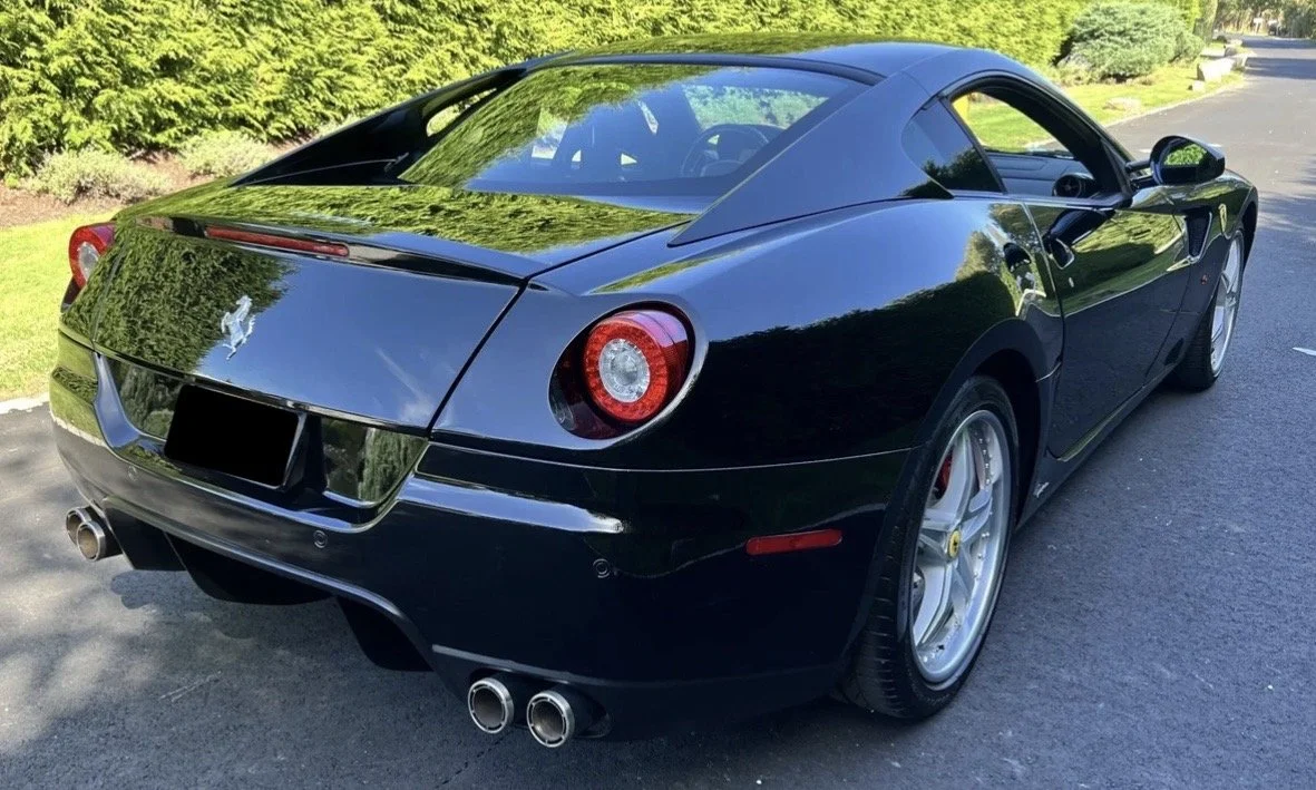 Black Ferrari sports car parked on a paved road with green trees and shrubs in the background.