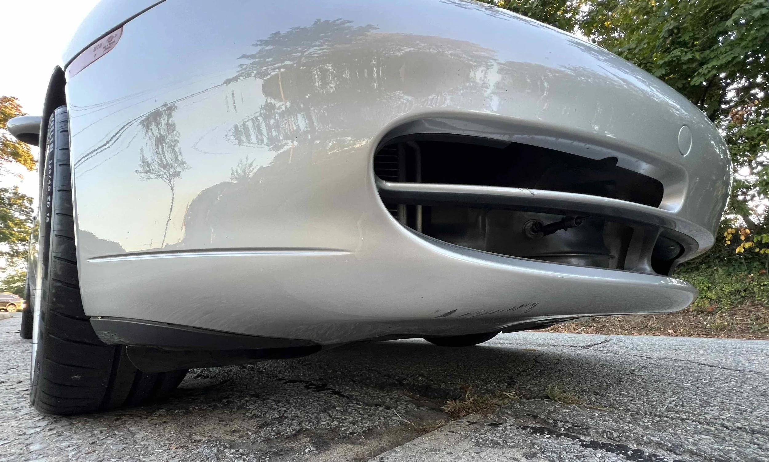 Close-up of the front part of a silver sports car, showing the front bumper and tire, parked on a paved surface with trees in the background.