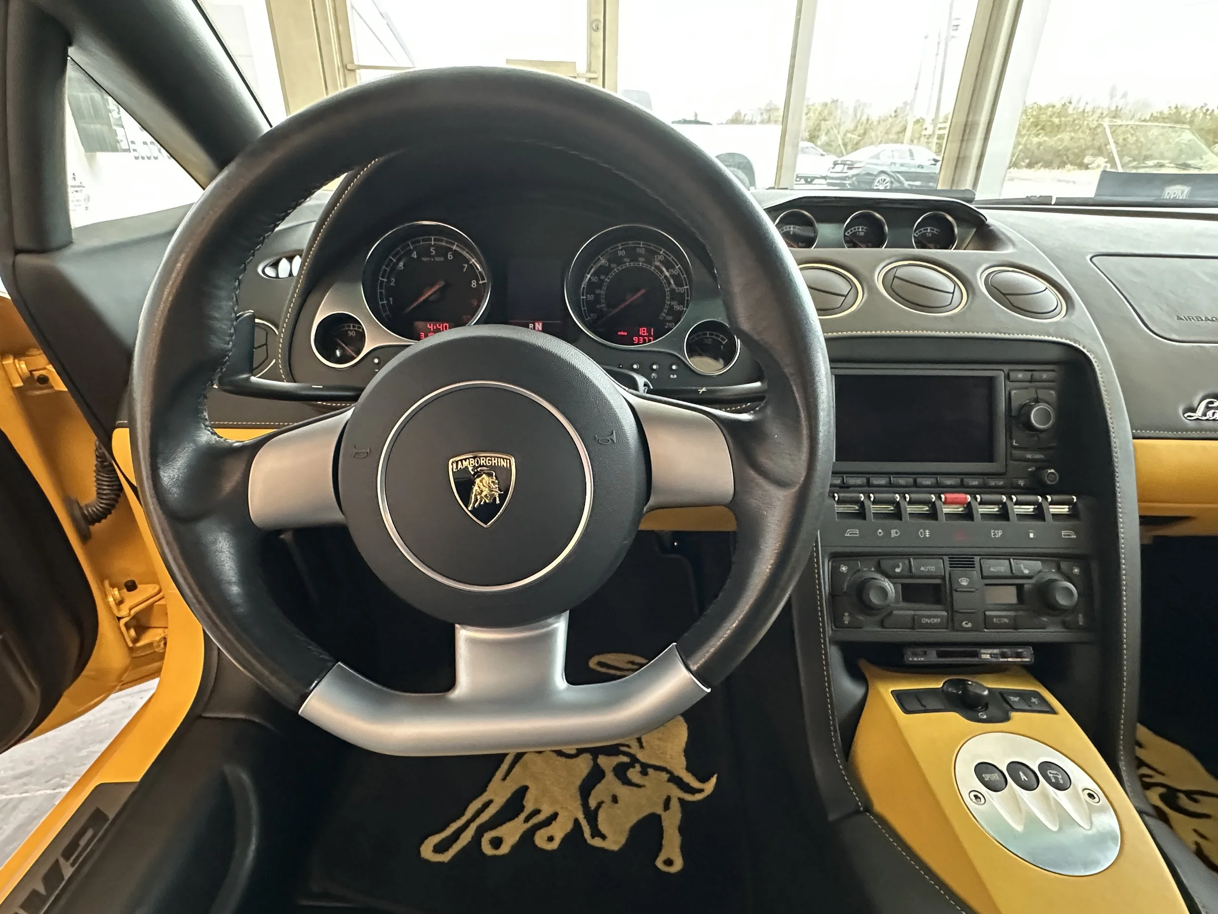 Interior of a Lamborghini sports car showing the steering wheel with Lamborghini logo, dashboard with speedometer and tachometer, center console with controls, and yellow accents with logo on floor mat.