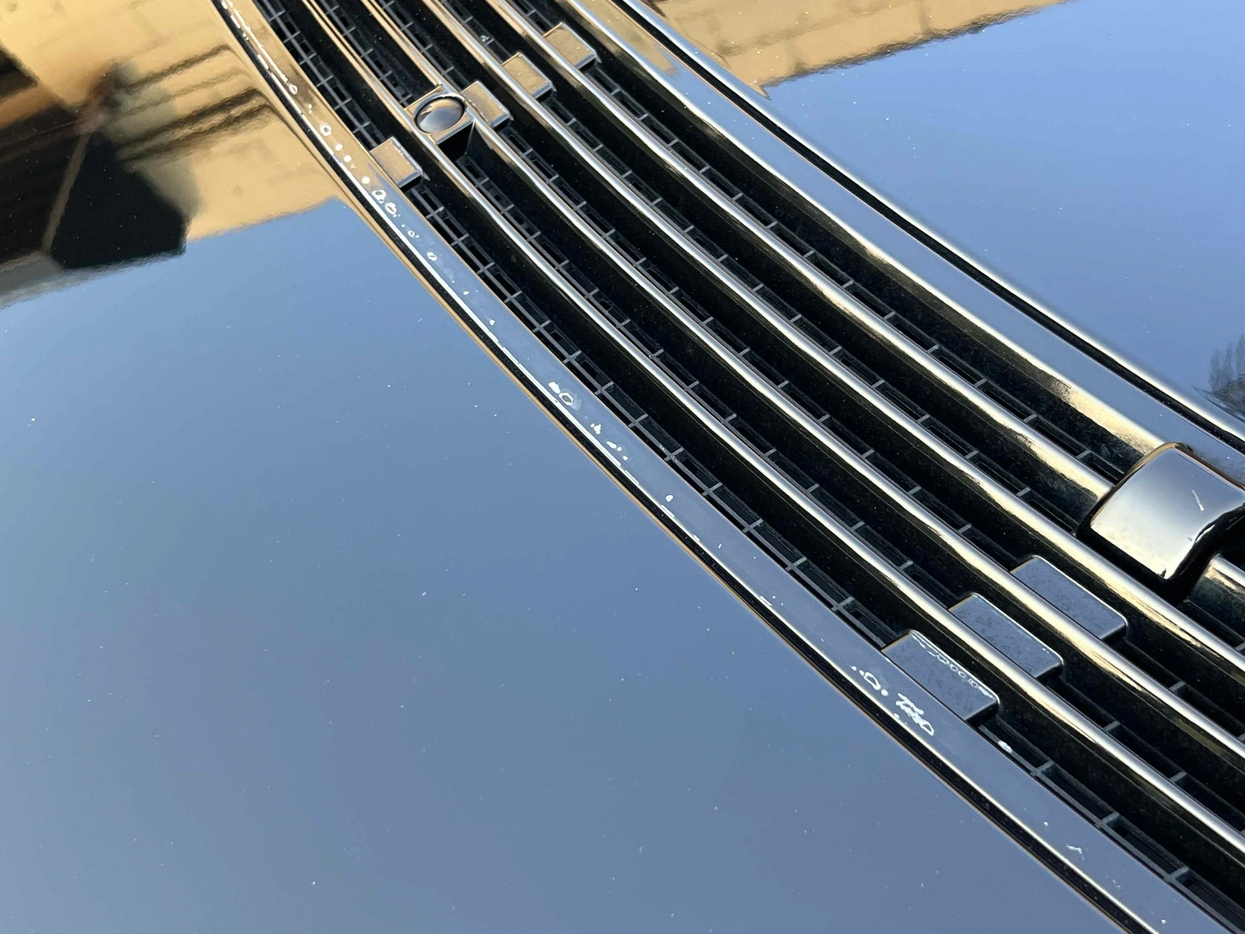 Close-up of a black car’s front grille and hood, with a reflection of the sky and a building on the shiny surface.