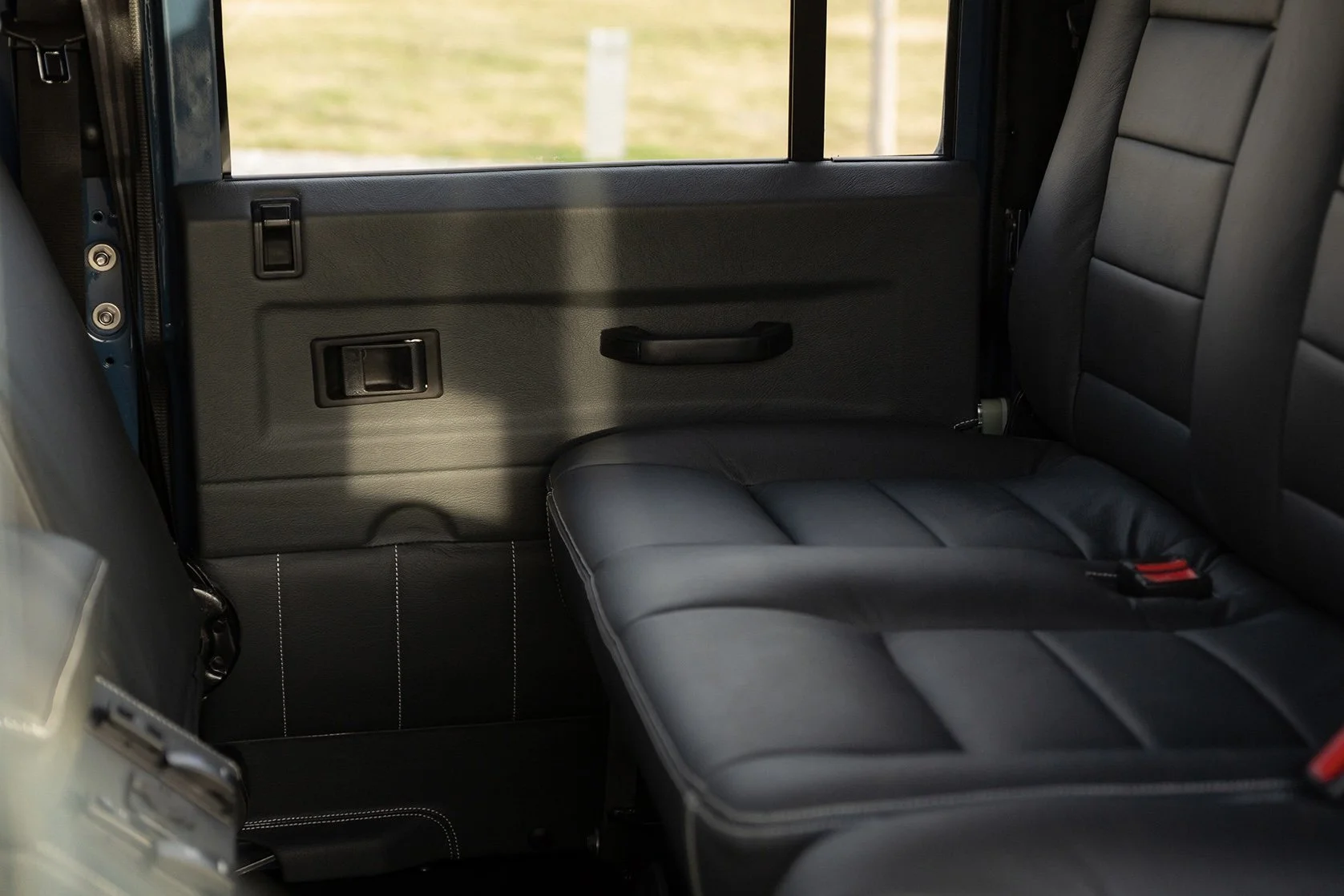 The interior of a vehicle showing a black leather backseat, a side door with a window, and a window view of an outdoor scene with grass.