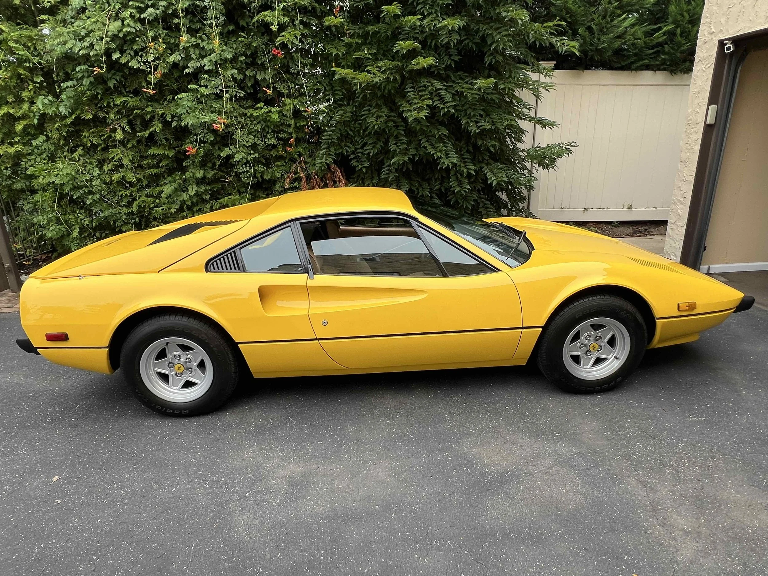 A yellow classic sports car parked on a driveway with a background of green bushes.