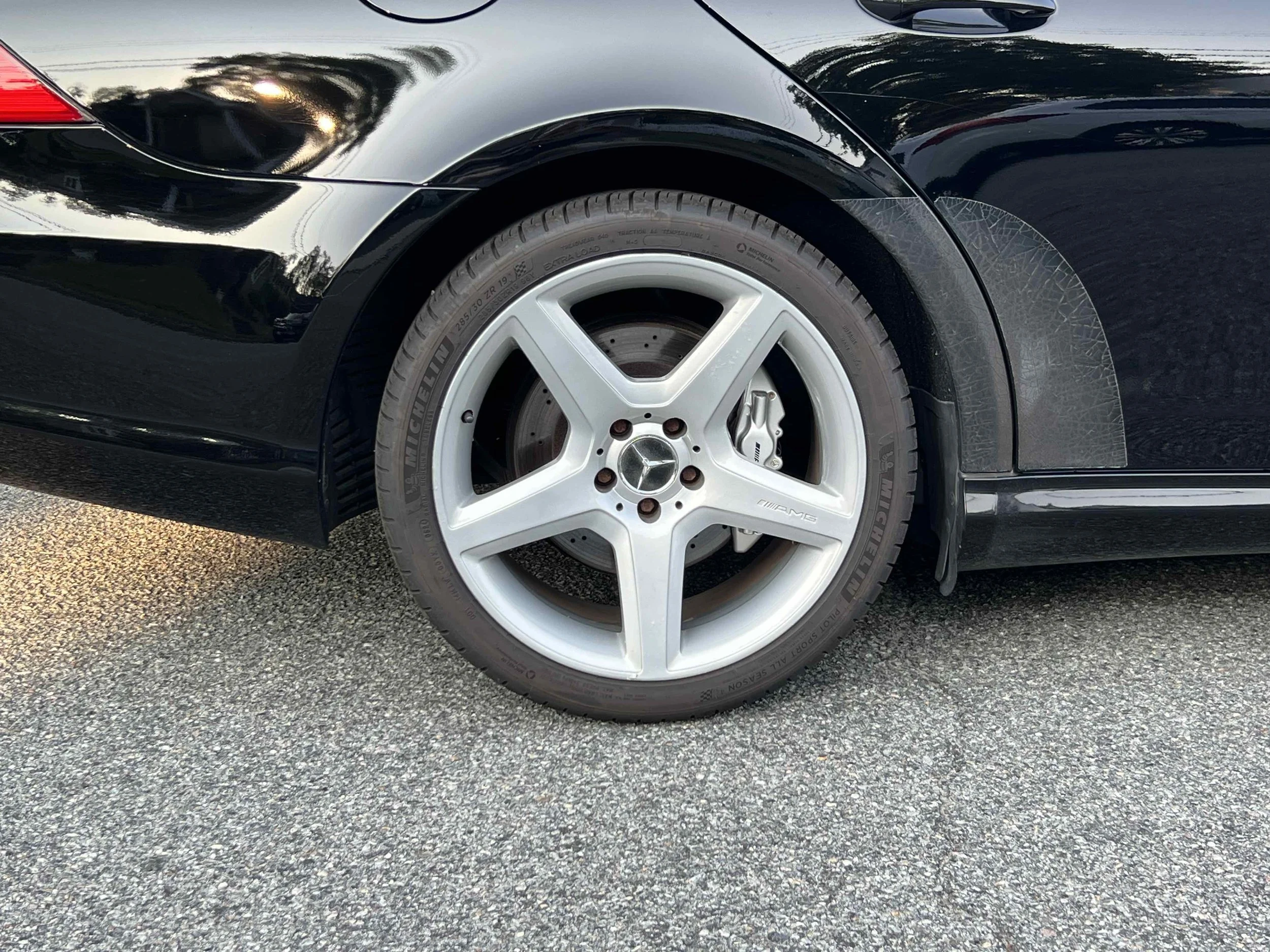 Close-up of a black Mercedes-Benz car wheel and tire, with the Mercedes logo visible on the hub, parked on an asphalt surface.