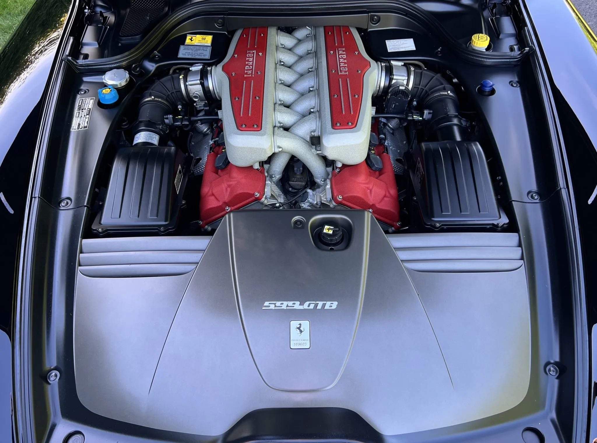 Under the hood of a Ferrari 599 GTB, showing the engine with red valve covers, metallic intake manifold, and black surrounding components.