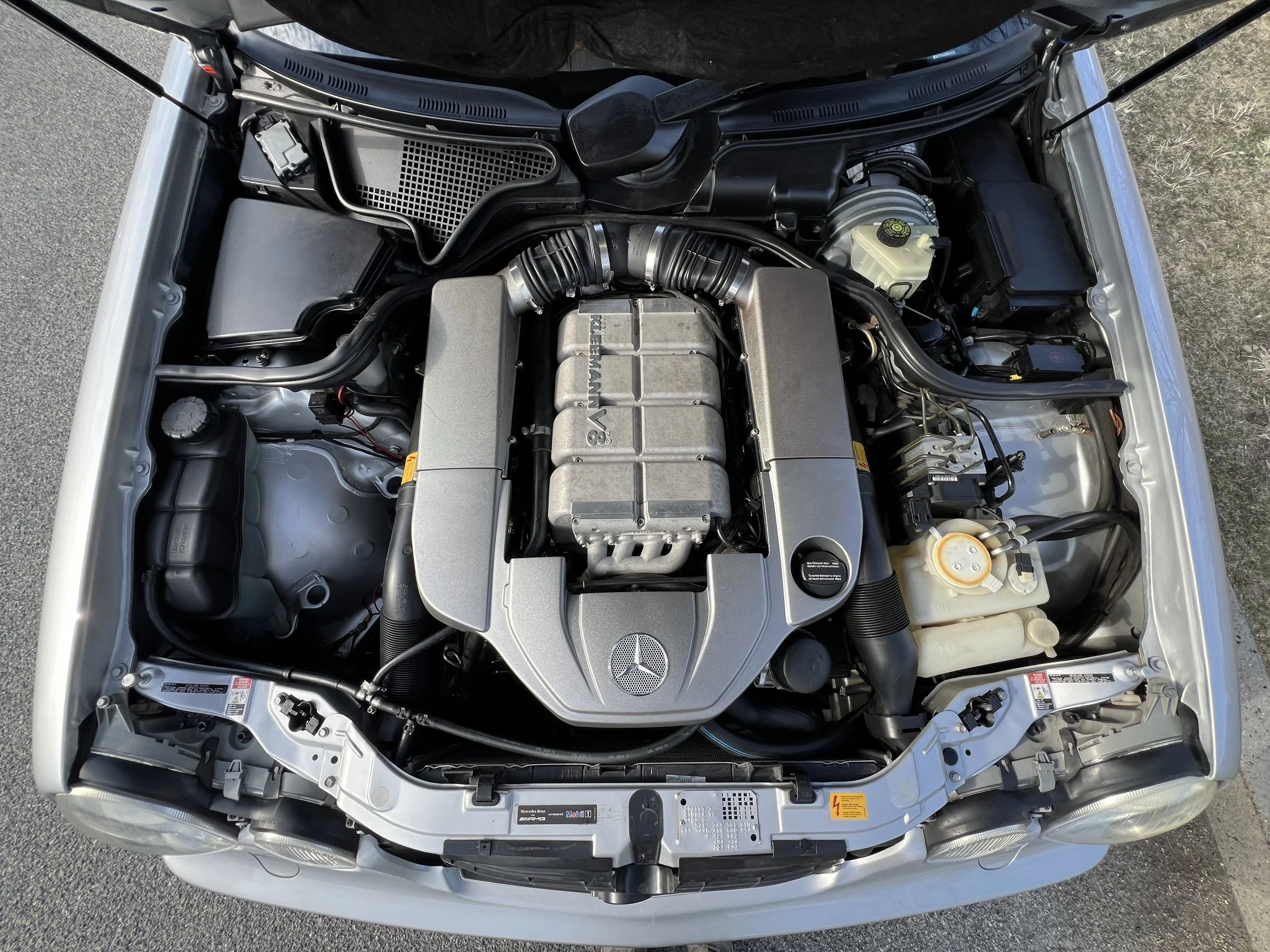 Engine bay of a silver car with visible Mercedes-Benz and AMG logos, showcasing various engine components, hoses, and fluid reservoirs.