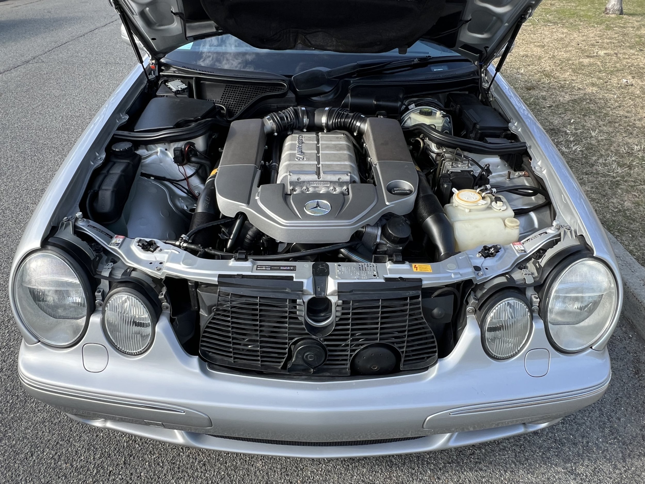 Front view of a silver sports car with its hood open, revealing the engine inside, parked on a road with grass on the side.