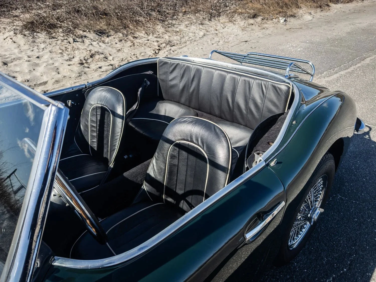 A vintage black convertible car parked on a paved area near a sandy and grassy terrain, showing the interior with black leather seats and chrome accents.