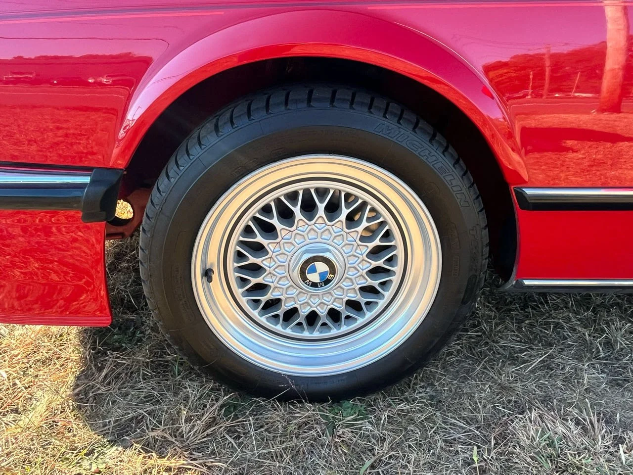 Close-up of a red car's wheel with a multi-spoke alloy rim and a BMW logo in the center, parked on a grassy area.