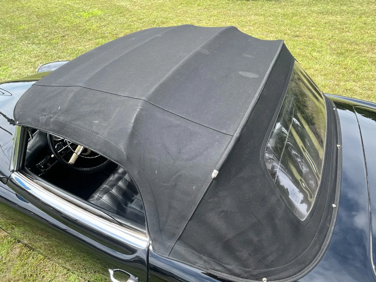 Black convertible car with soft top roof parked on grass, showing the interior steering wheel and front seats, with a clear rear windshield.