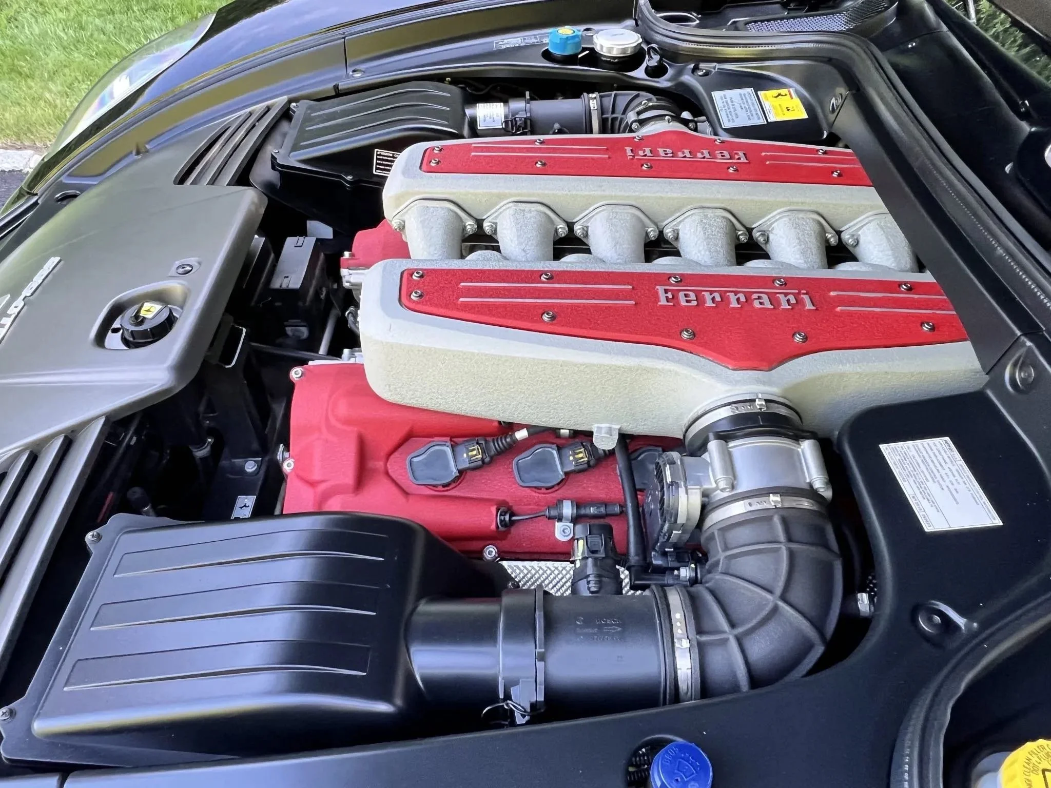 Close-up of a Ferrari engine bay with red and silver engine covers, black intake components, and various hoses and connectors.