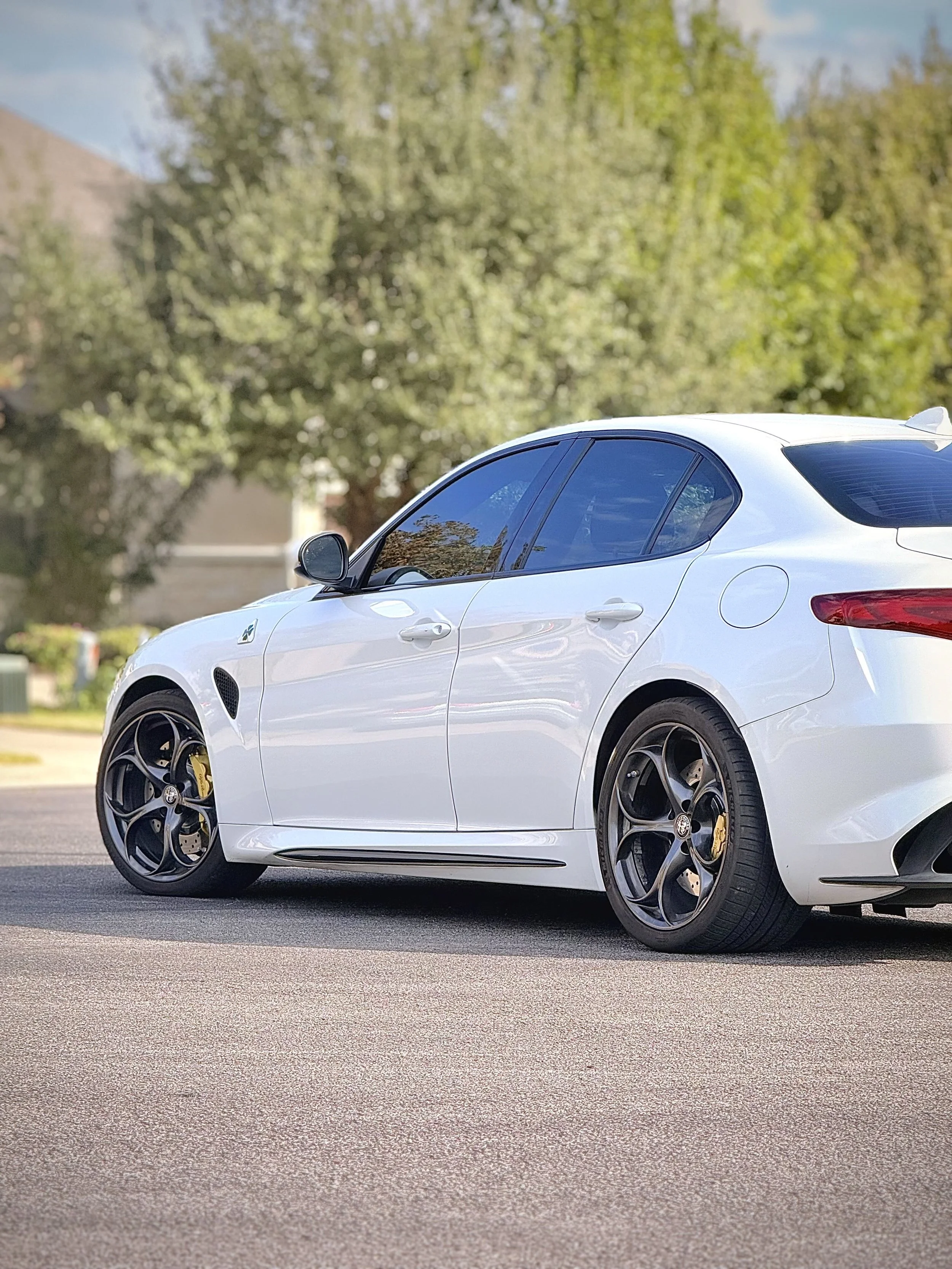 A white sports hatchback car parked on an asphalt street with green trees and a house in the background.