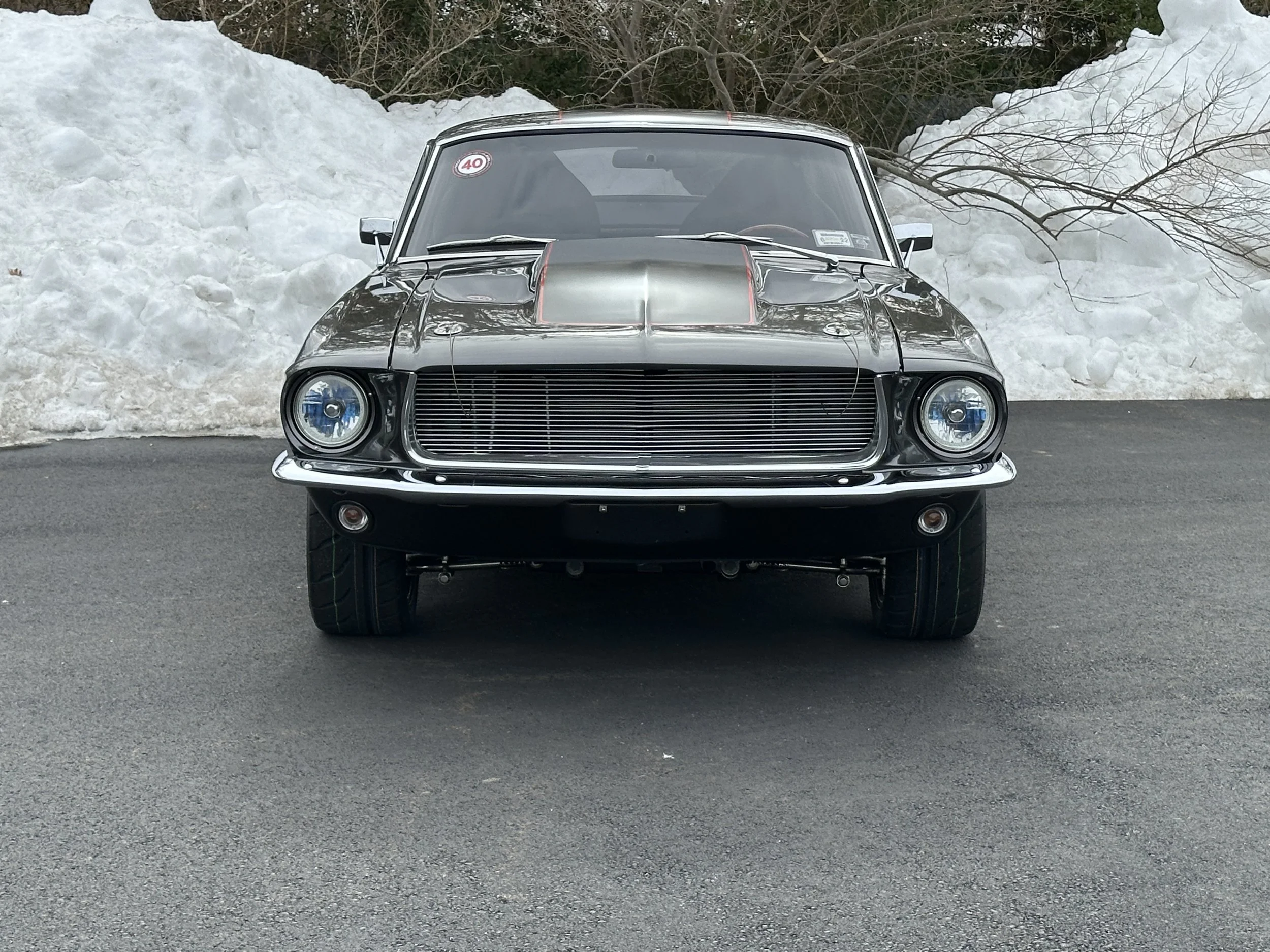 Front view of a classic black muscle car with a custom grille and hood, parked on asphalt with snow piles and trees in the background.