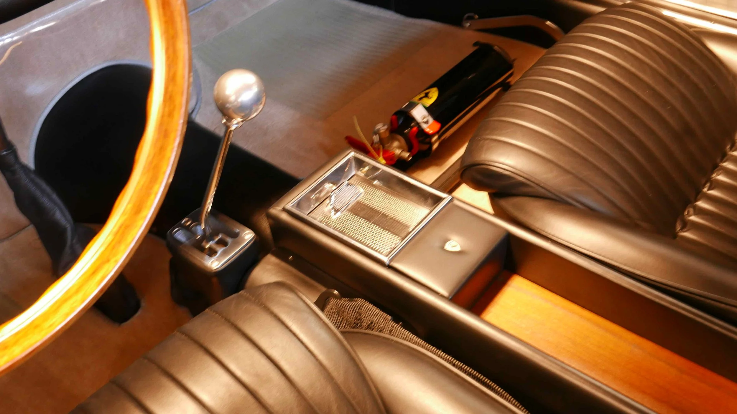 Inside a classic car with black leather seats, gear shifter, cigarette lighter, and small luggage or bag on the side.