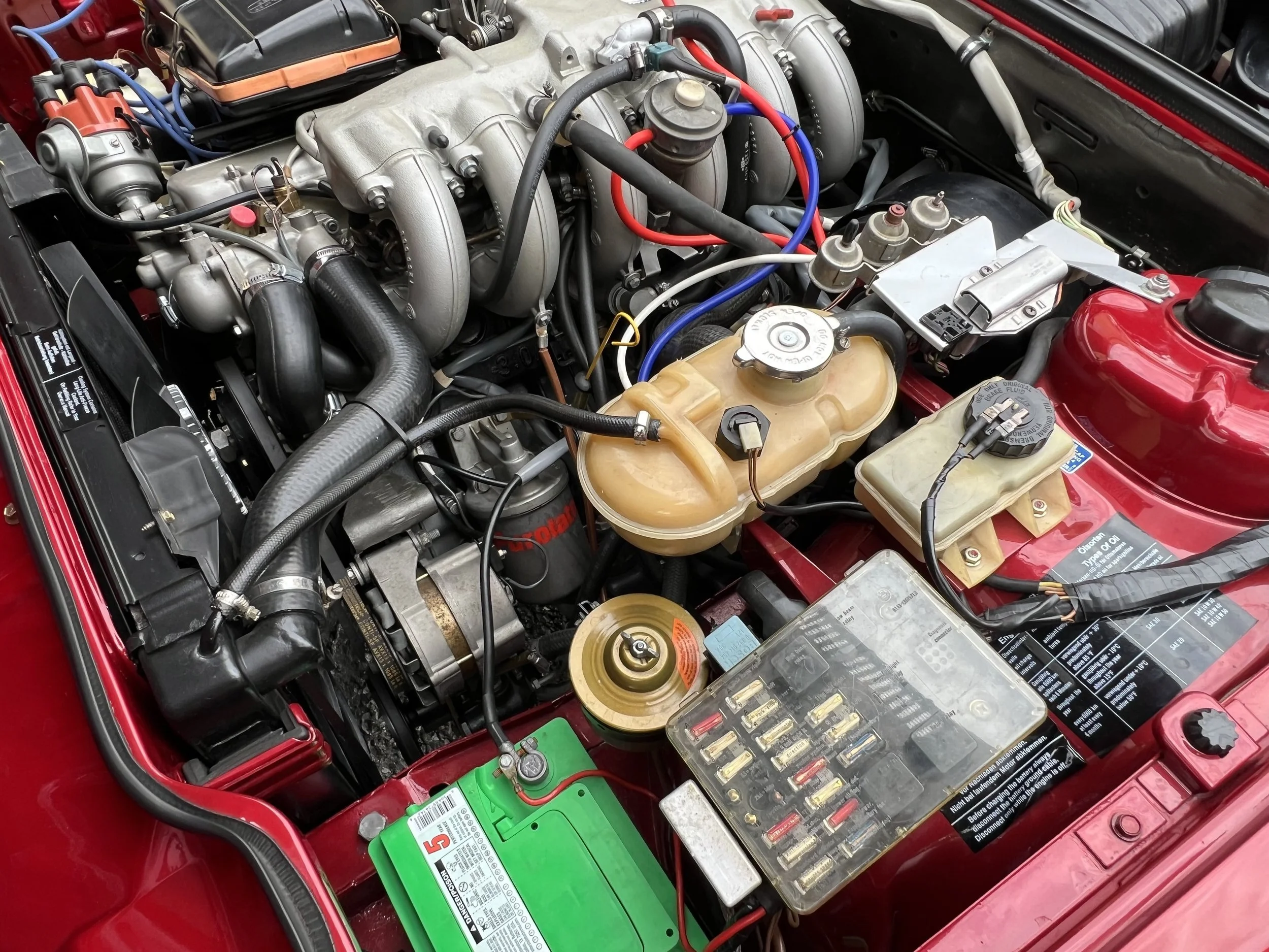 Close-up of a 1977 BMW 630 csi car engine bay showing various mechanical and electrical components, including the battery, fuse box, coolant reservoir, and intake manifold.
