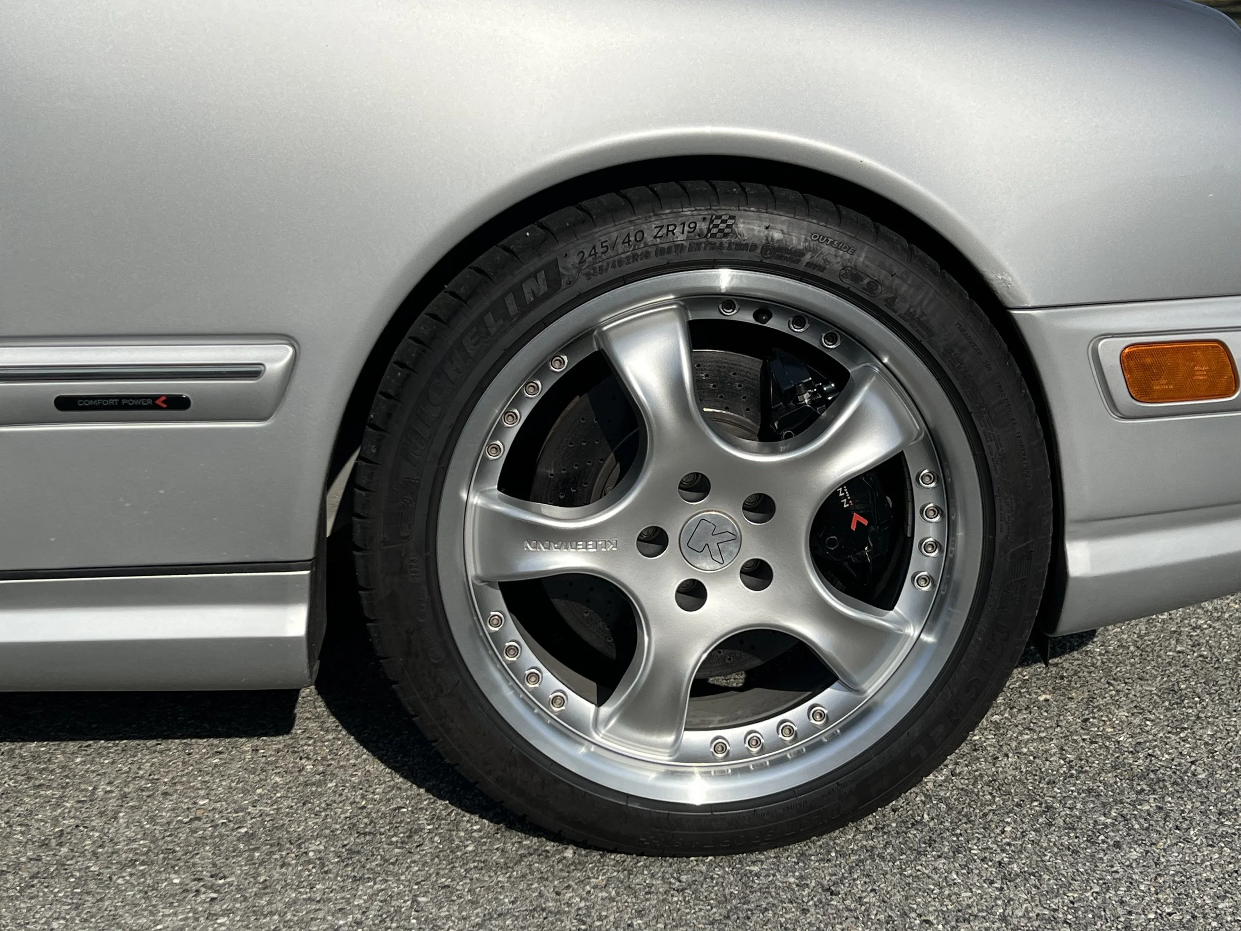 Close-up of a silver car's front wheel with a sports tire and aftermarket rim, showing part of the car's fender, side marker light, and the ground.