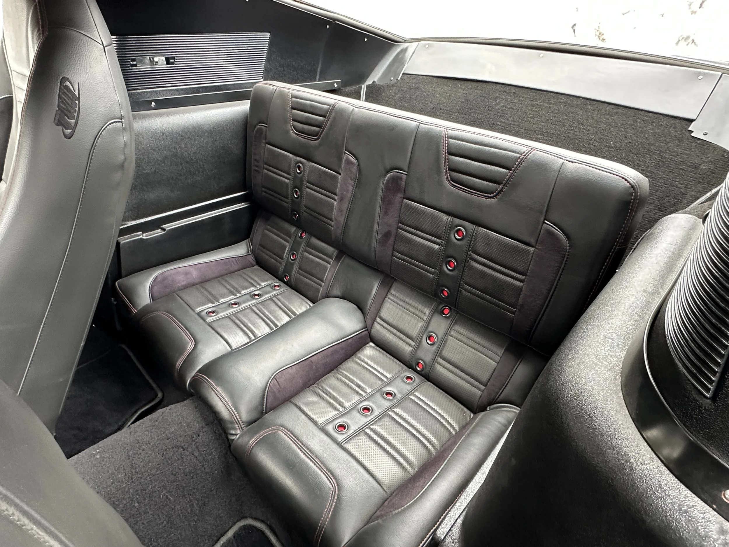 The image shows the interior of a vintage car, featuring a black leather rear bench seat with red stitching and button accents, and other surrounding interior details.