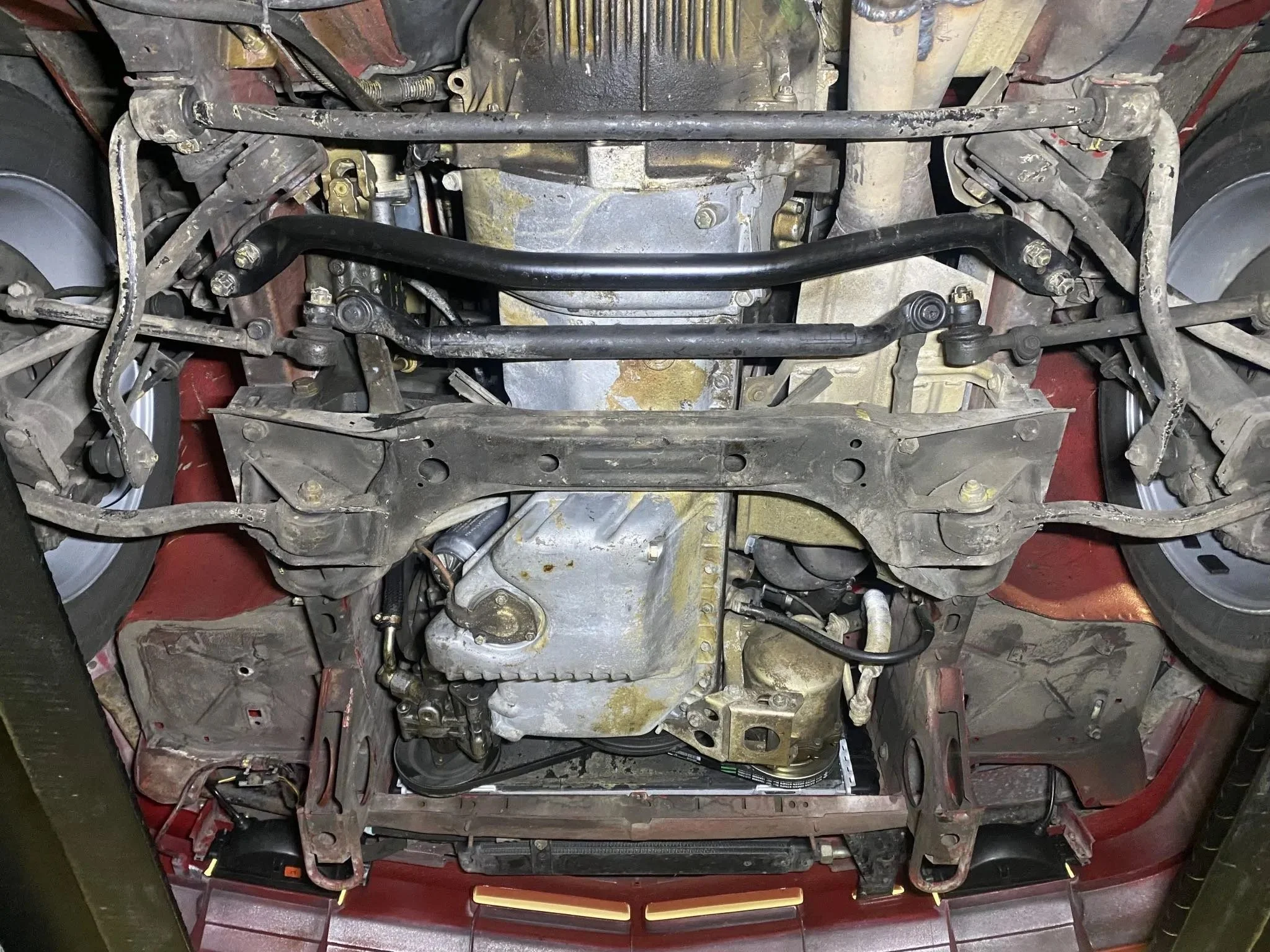 Under the car's chassis showing suspension components, steering linkages, and engine parts, including the oil pan and surrounding metal framework.
