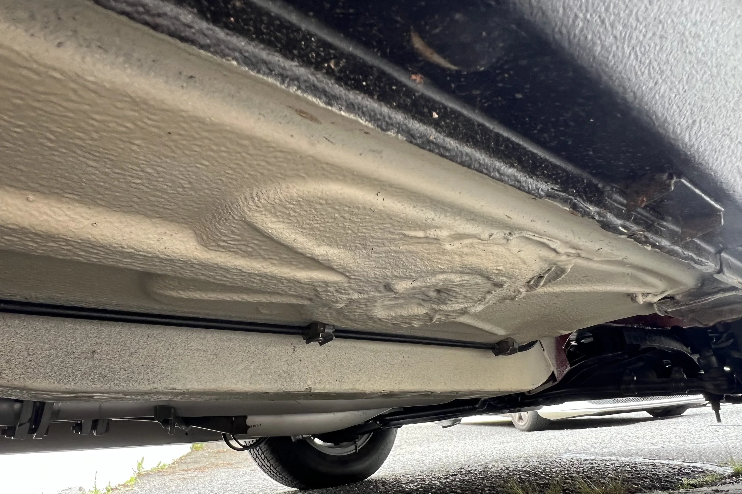 Underside of a 1977 BMW 630 csi vehicle showing the beige undercoating and part of the exhaust system.
