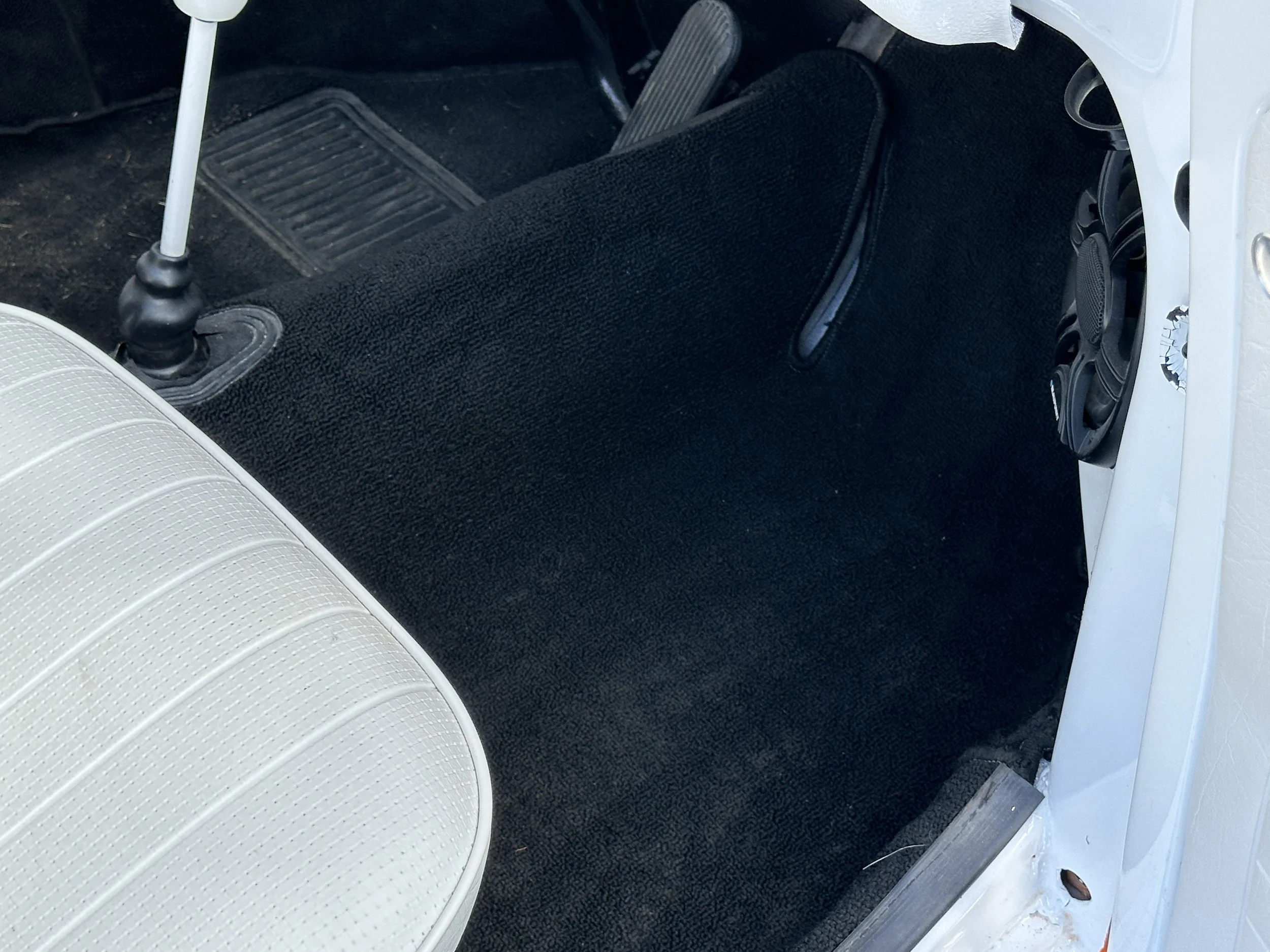 The interior of a vintage car showing the black carpeted floor, the edge of a white seat, and the lower part of a gear shifter.