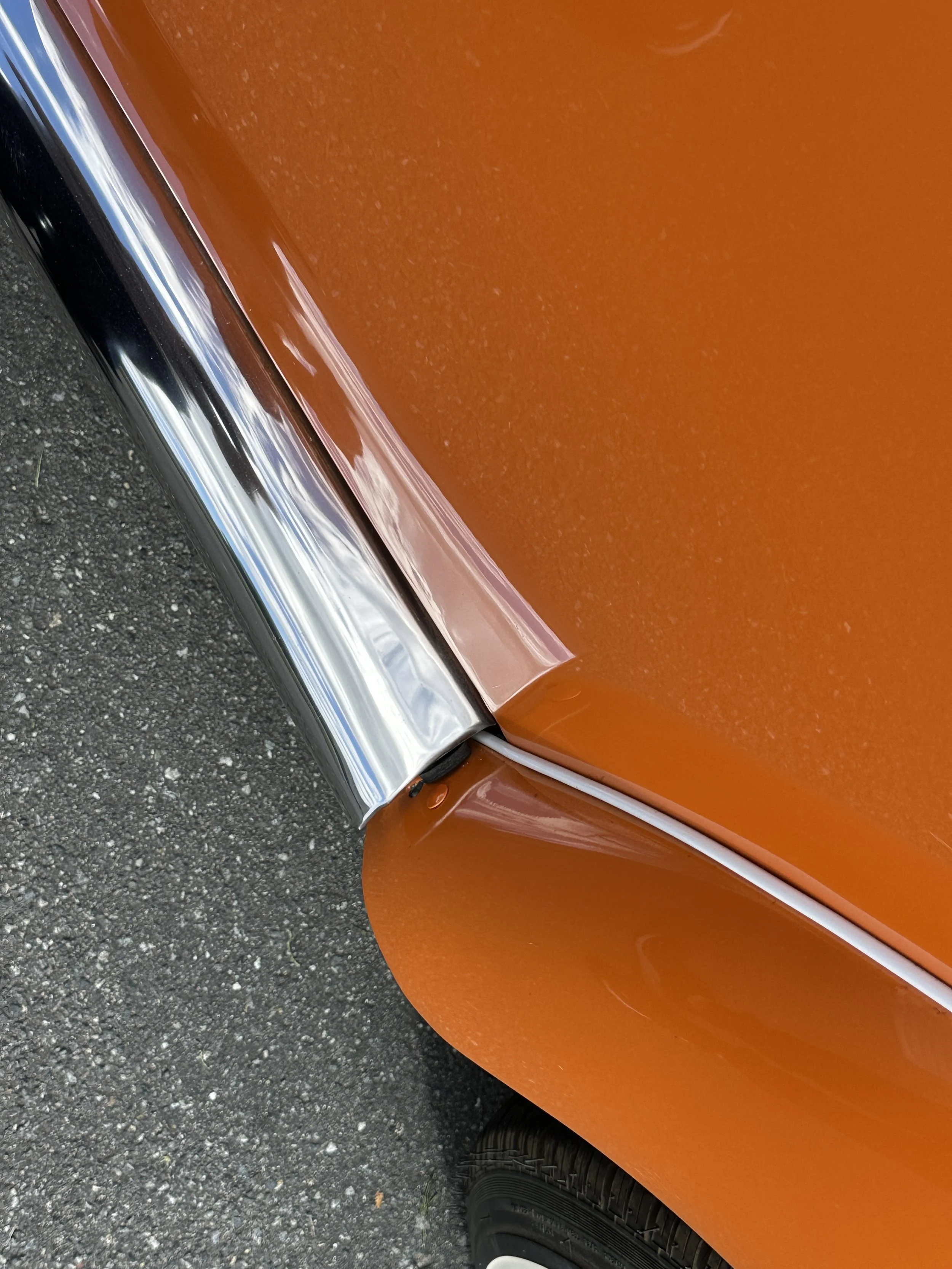 Close-up of a vintage orange car's wheel well, chrome trim, and part of the tire on asphalt pavement.