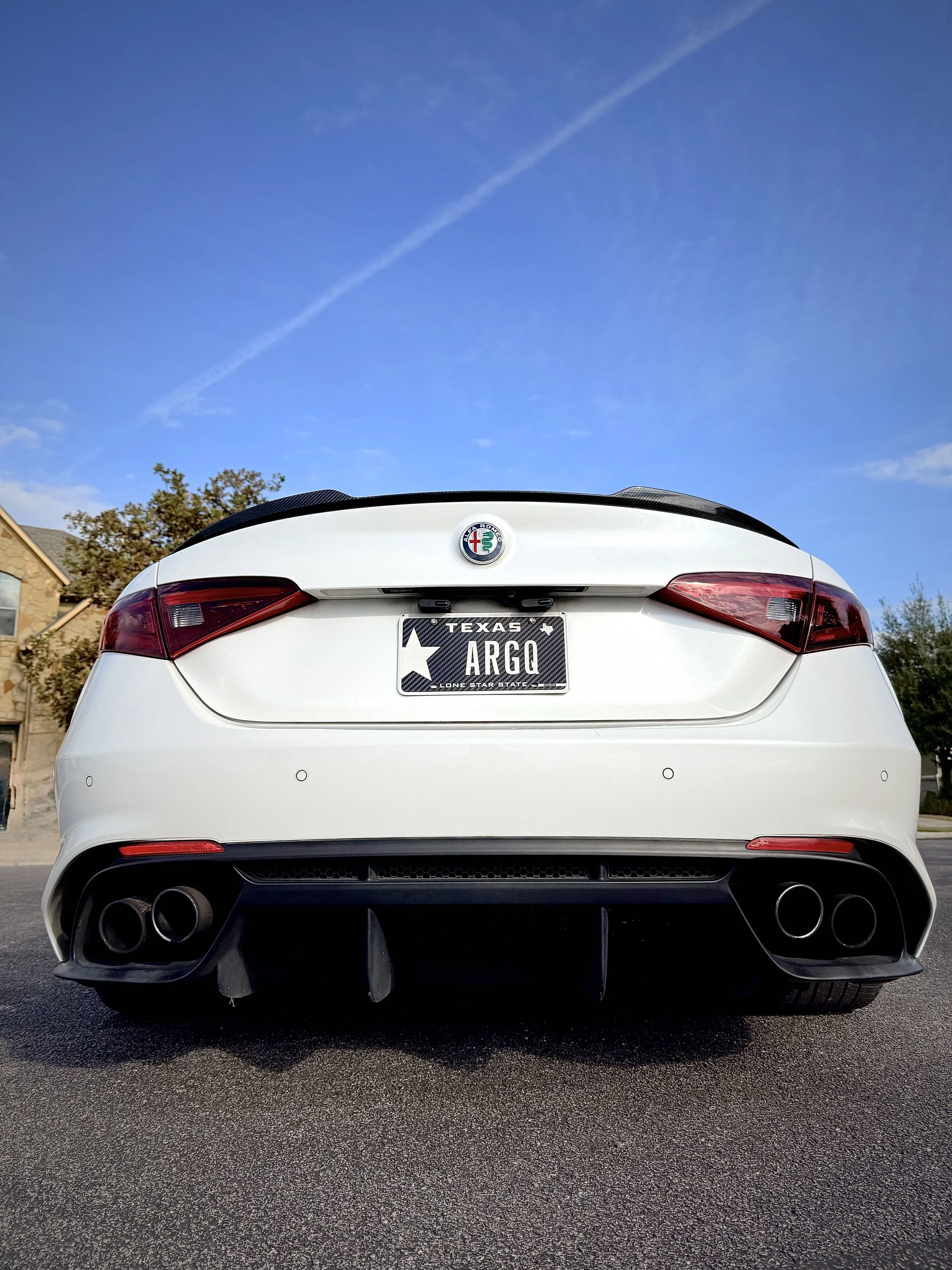 Rear view of a white Alfa Romeo car with Texas license plate reading 'ARGO', parked on a street under a blue sky.