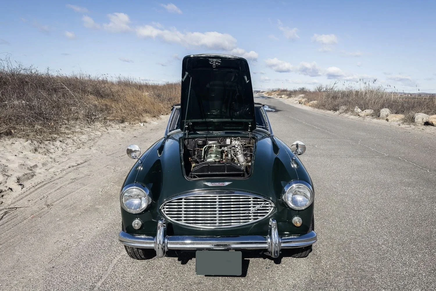 Front view of a vintage dark green sports car with its hood open, parked on a rural road with dry bushes on sides under a partly cloudy sky.