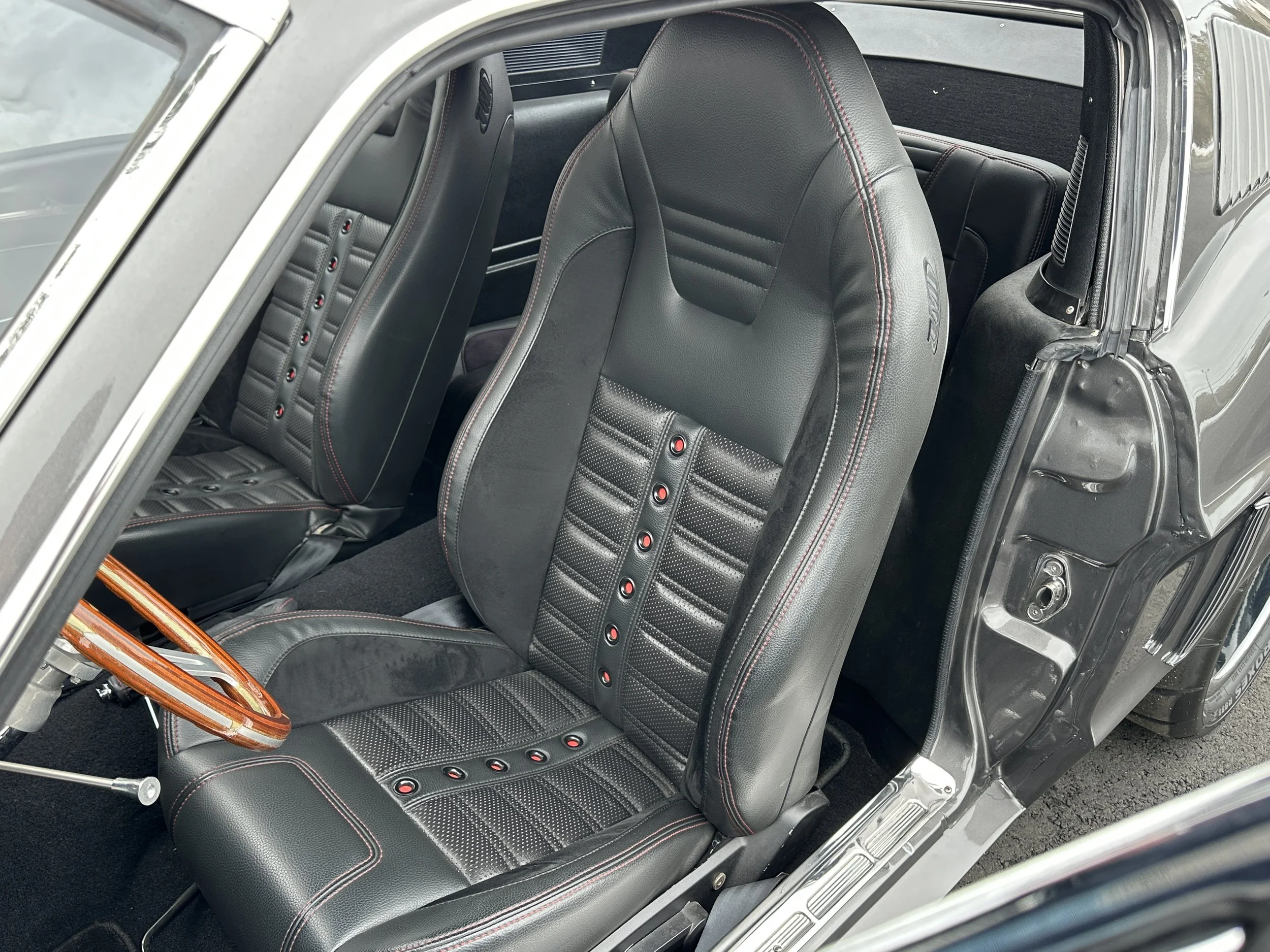 The interior of a classic sports car featuring black leather seats with red stitching and sporty design, a wooden steering wheel, and a minimalist dashboard.
