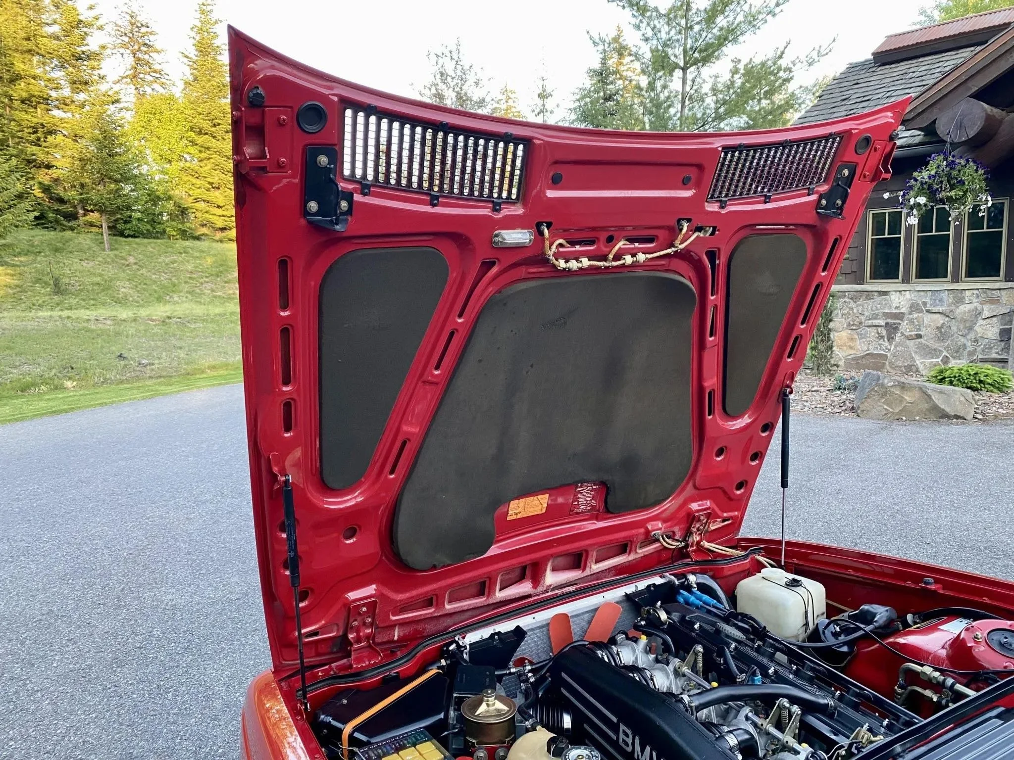 View of the hood of a red sports car with engine components visible, set outdoors with a house and trees in the background.