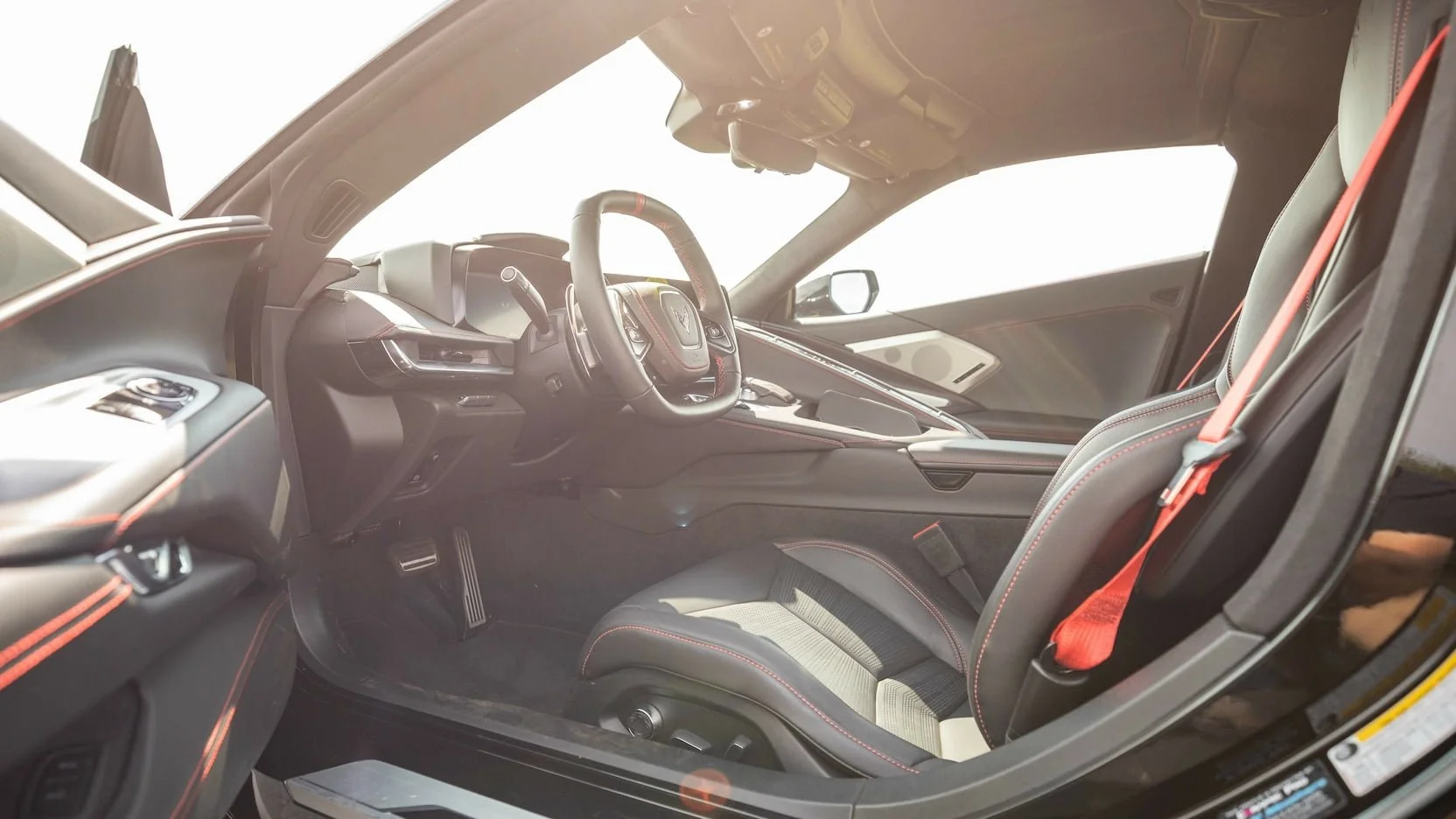 The interior of a modern sports car showing the driver's seat, steering wheel, dashboard, and front passenger seat, with black and red accents.
