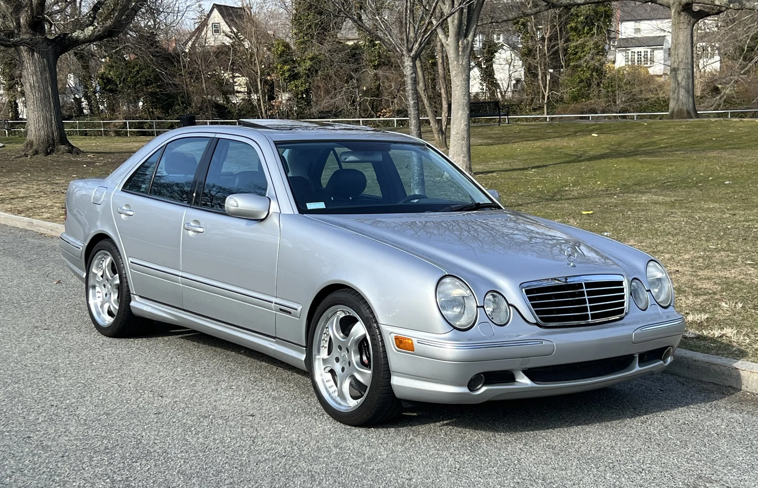 A silver Mercedes-Benz sedan parked on the side of a street with trees and grass in the background.