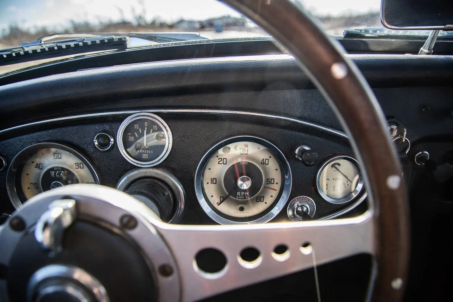 Dashboard of a vintage car with speedometer, tachometer, amperes gauge, and fuel gauge, viewed from the driver's seat with a wooden steering wheel.
