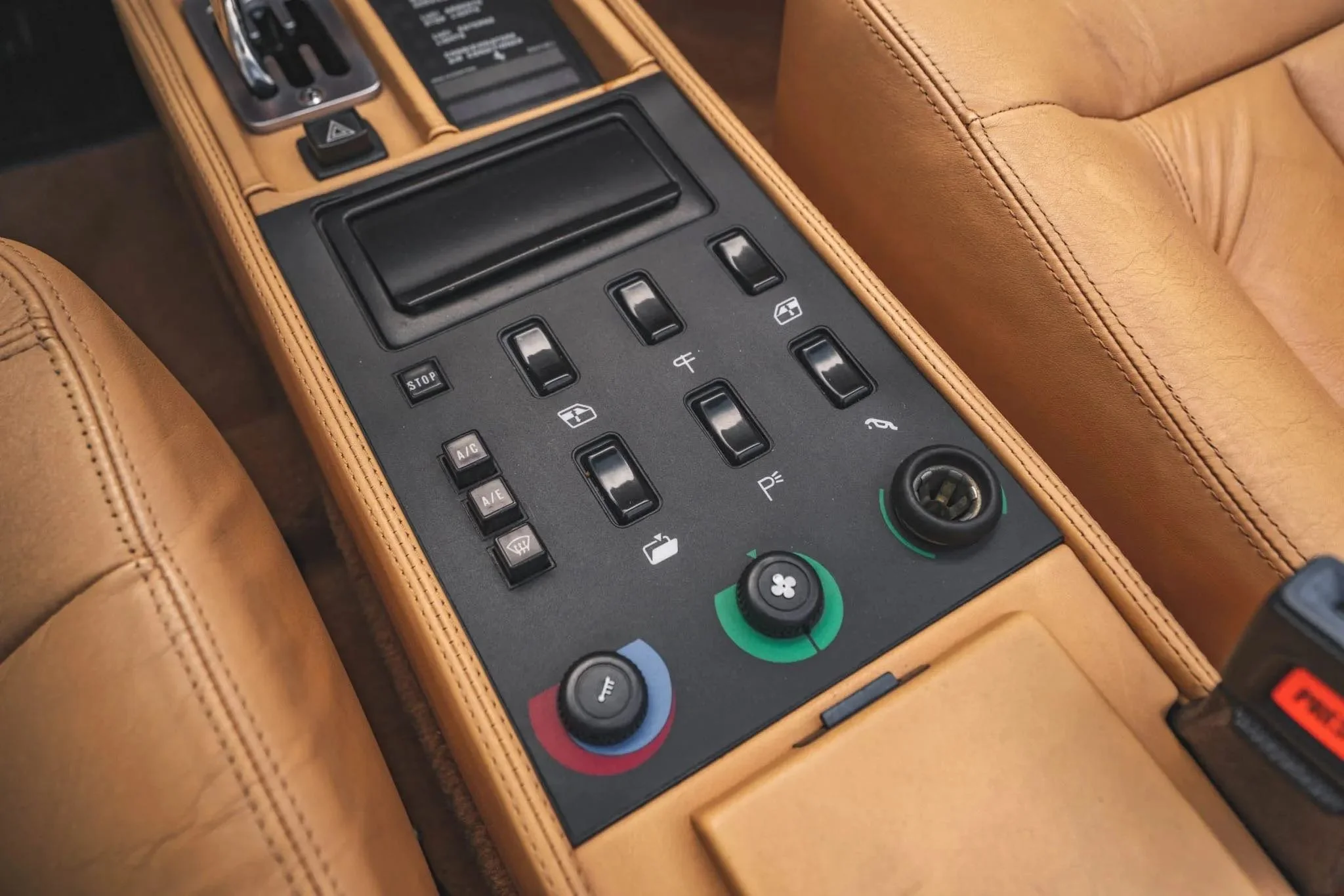 Close-up of a car's center console with tan leather upholstery, featuring various climate control switches, buttons, and dials for adjusting temperature, fan speed, and airflow.