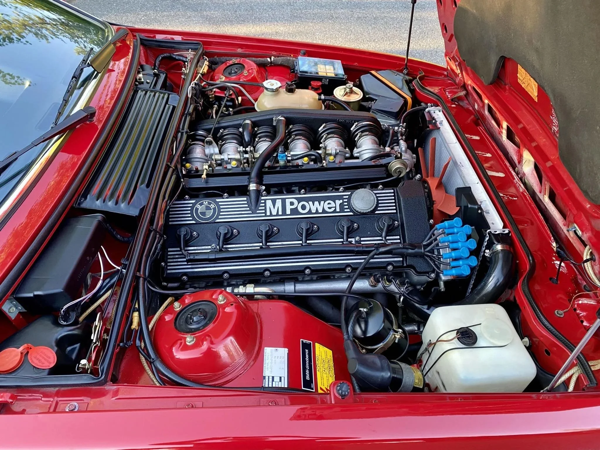 Under the open hood of a red BMW, the engine bay features a black M Power engine cover, visible intake manifold, and various engine components including hoses, wires, and the coolant reservoir.