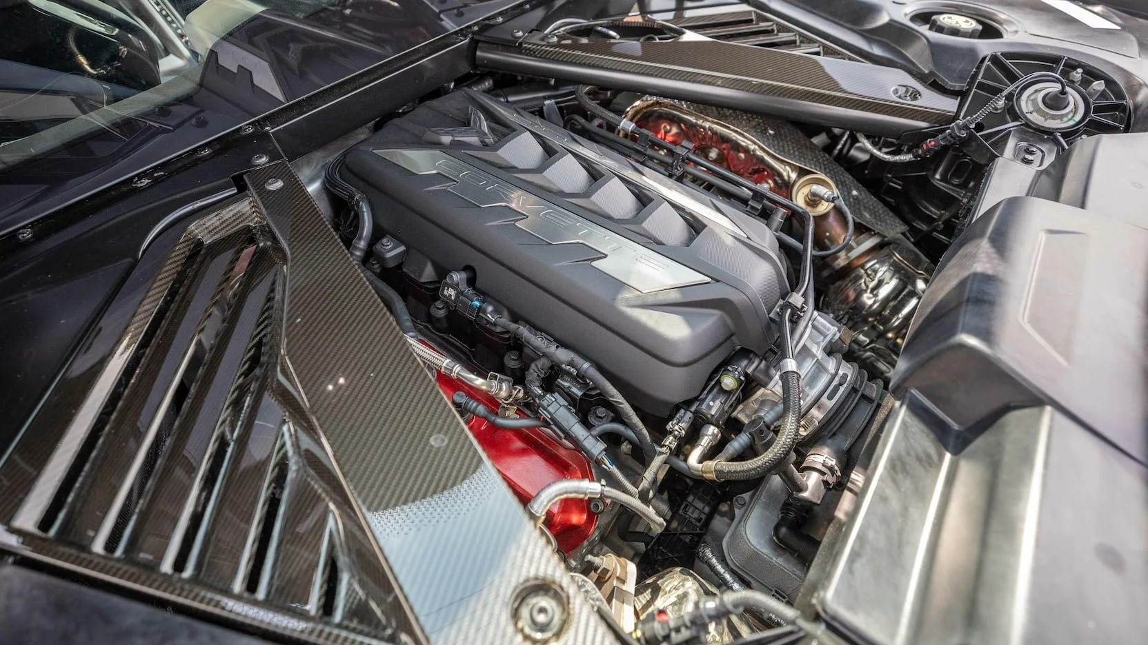 Close-up of a car engine bay featuring a carbon fiber hood vent, a black engine cover with the word 'Corvette' embossed, and various engine components and hoses.