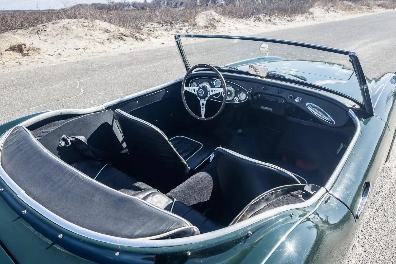 Interior of a vintage convertible car parked on a dirt road with a sandy hill in the background, showing the steering wheel and dashboard.
