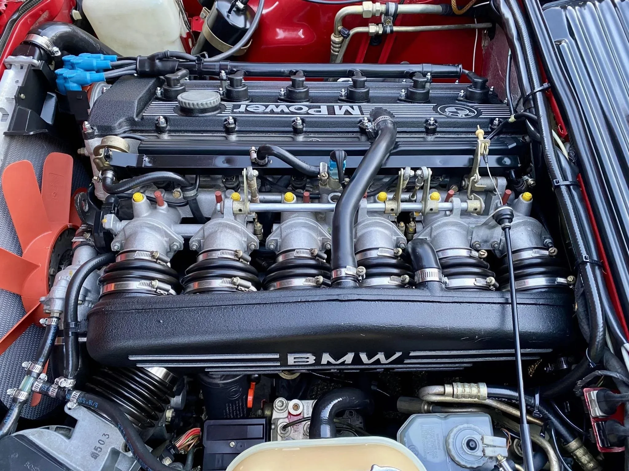 Close-up of a BMW engine with visible BMW logo on the valve cover, various hoses, and mechanical components.