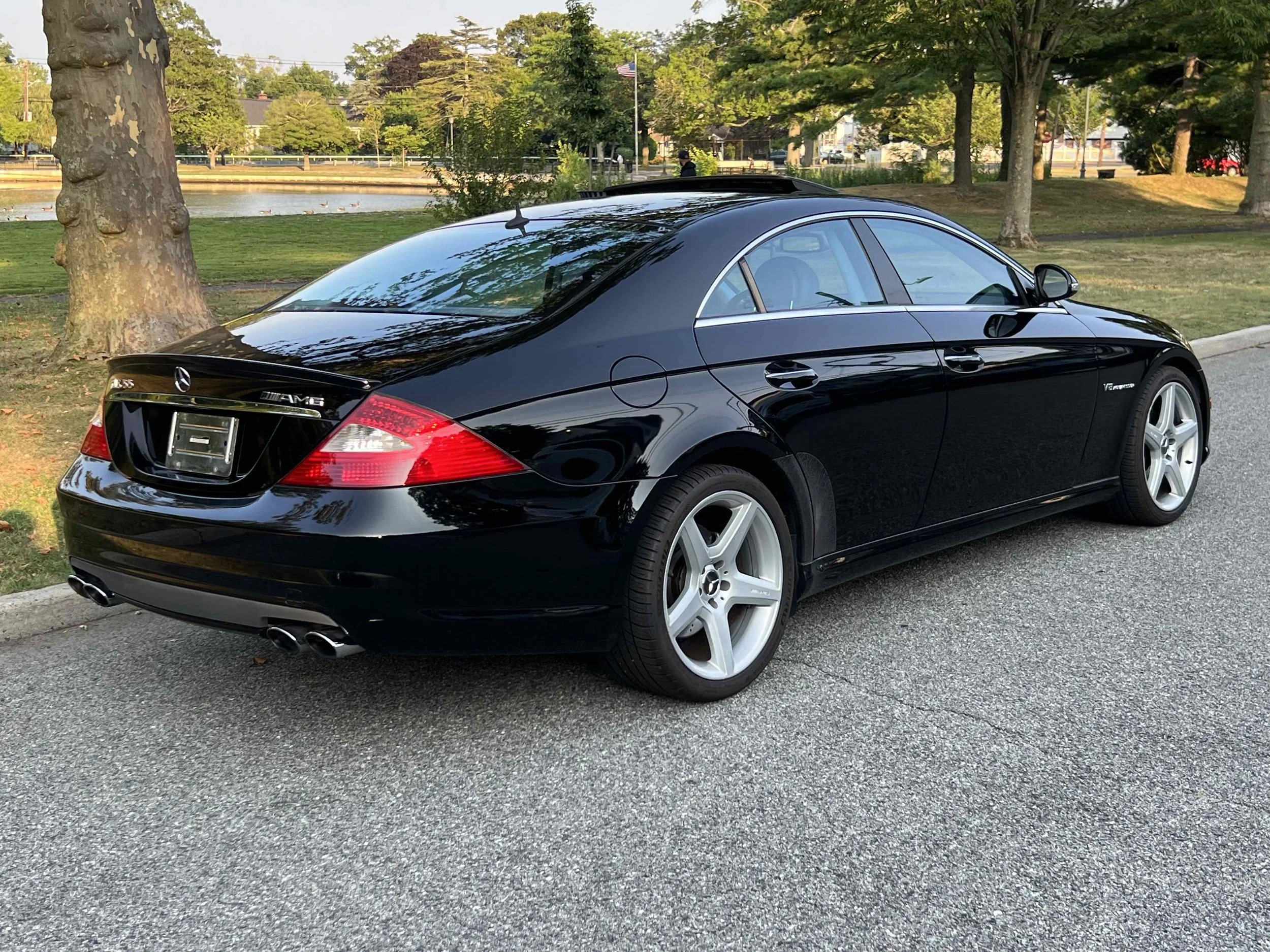Black Mercedes-Benz CLS55 AMG  parked on the side of a street with trees and a pond in the background.