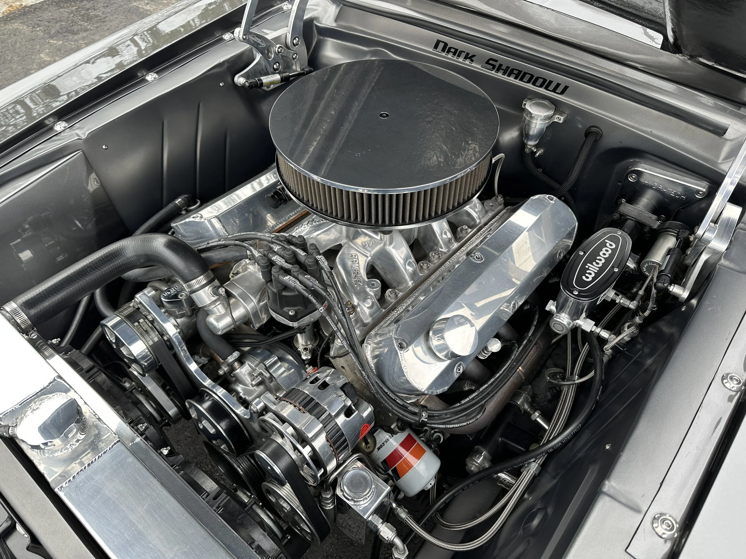 A high-performance car engine with a chrome valve cover, a round air cleaner, and Wilwood brake components.