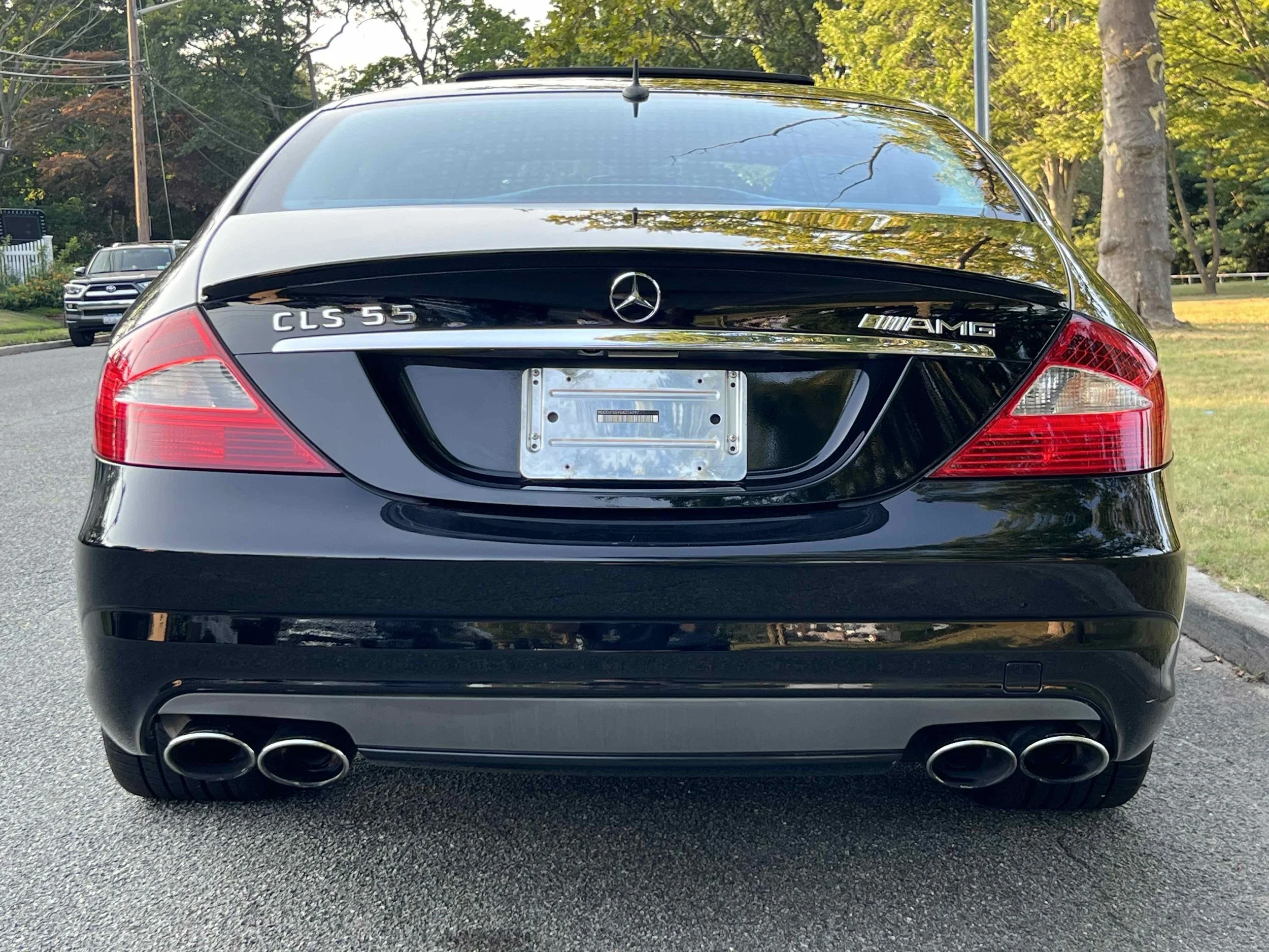 Rear view of a black Mercedes-Benz CLS 55 AMG, four exhaust pipes, and showroom taillights, parked on a suburban street with trees and other vehicles in the background.