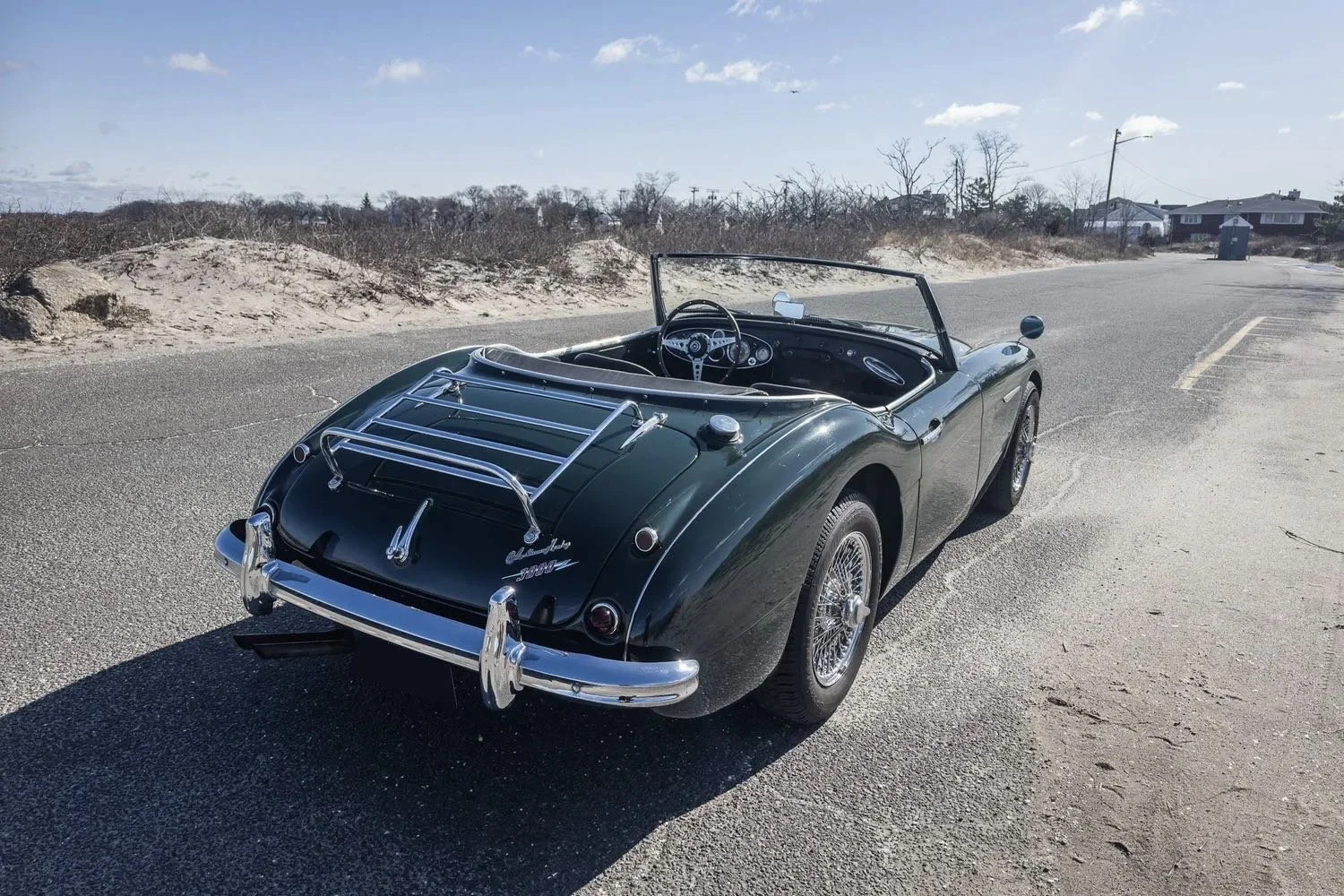 A vintage dark green convertible sports car parked on a coastal road with a sandy beach and beach dunes in the background, under a partly cloudy sky.
