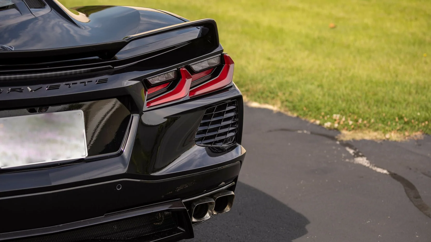 Close-up of the rear of a black Chevrolet Corvette, showing the taillights, exhaust pipes, and rear spoiler, parked on a paved surface with grass in the background.