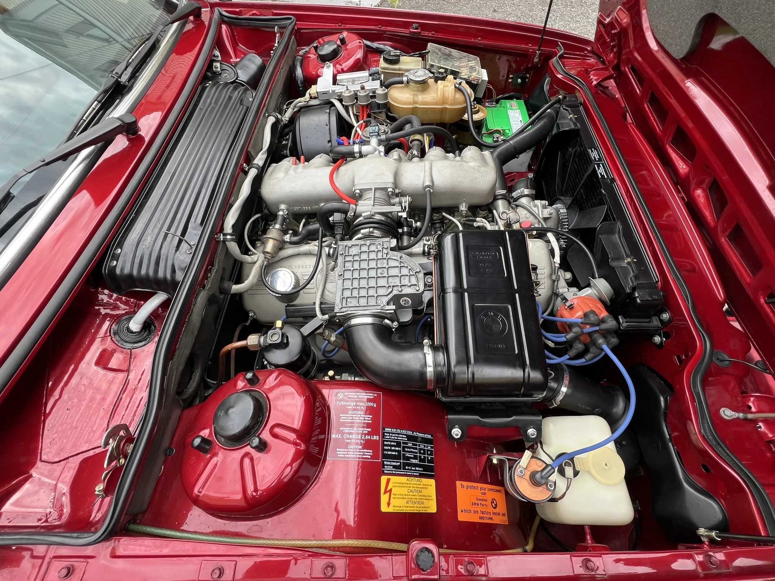 Red 1977 BMW 630 csi car engine bay with various components, including a large black air intake, orange and black battery, black engine cover, red coolant reservoir, and other engine parts.