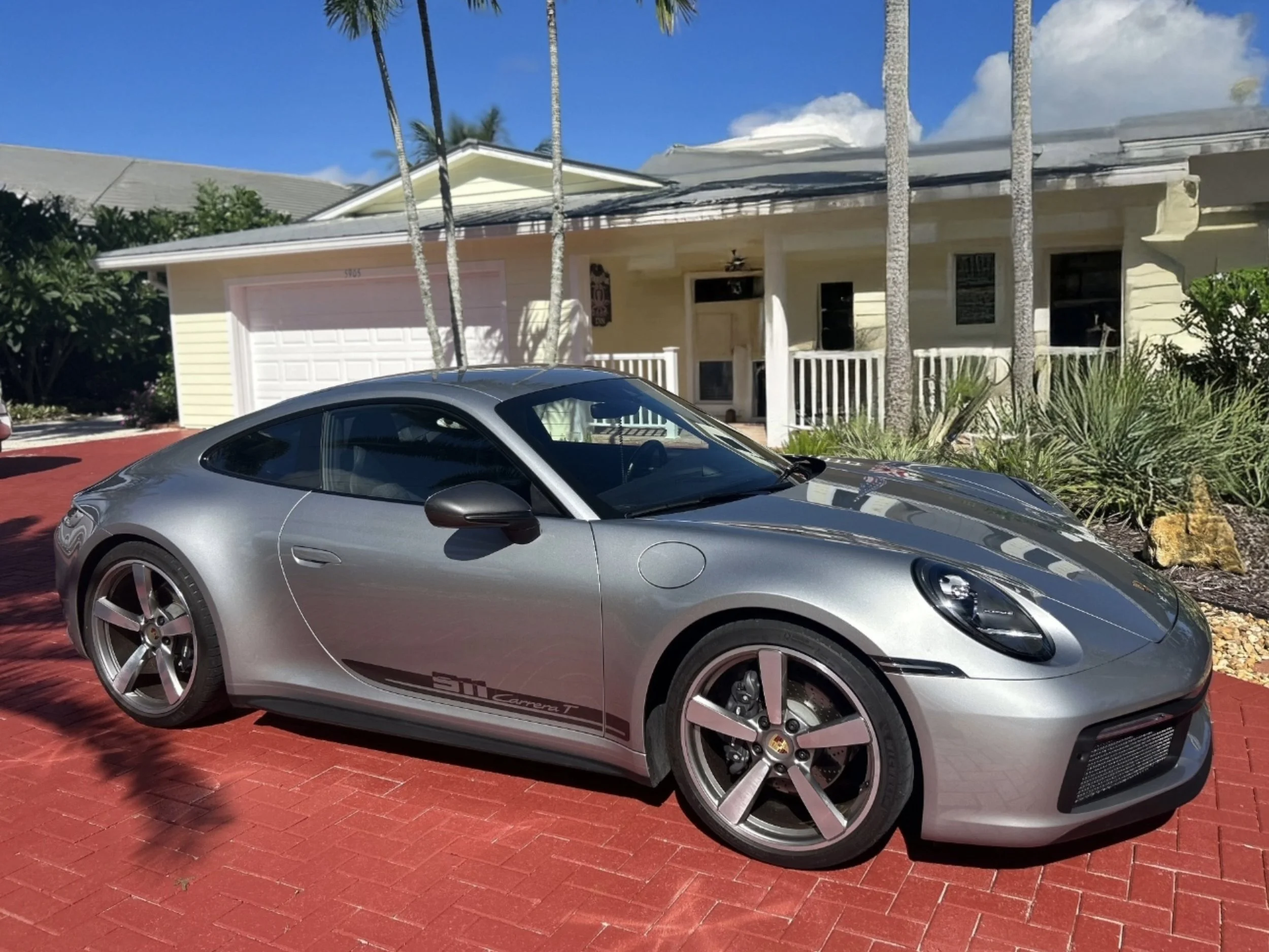 A silver Porsche 911 Carrera T parked on a red brick driveway in front of a yellow house with a white garage door and porch. Palm trees are in the background under a sunny, partly cloudy sky.